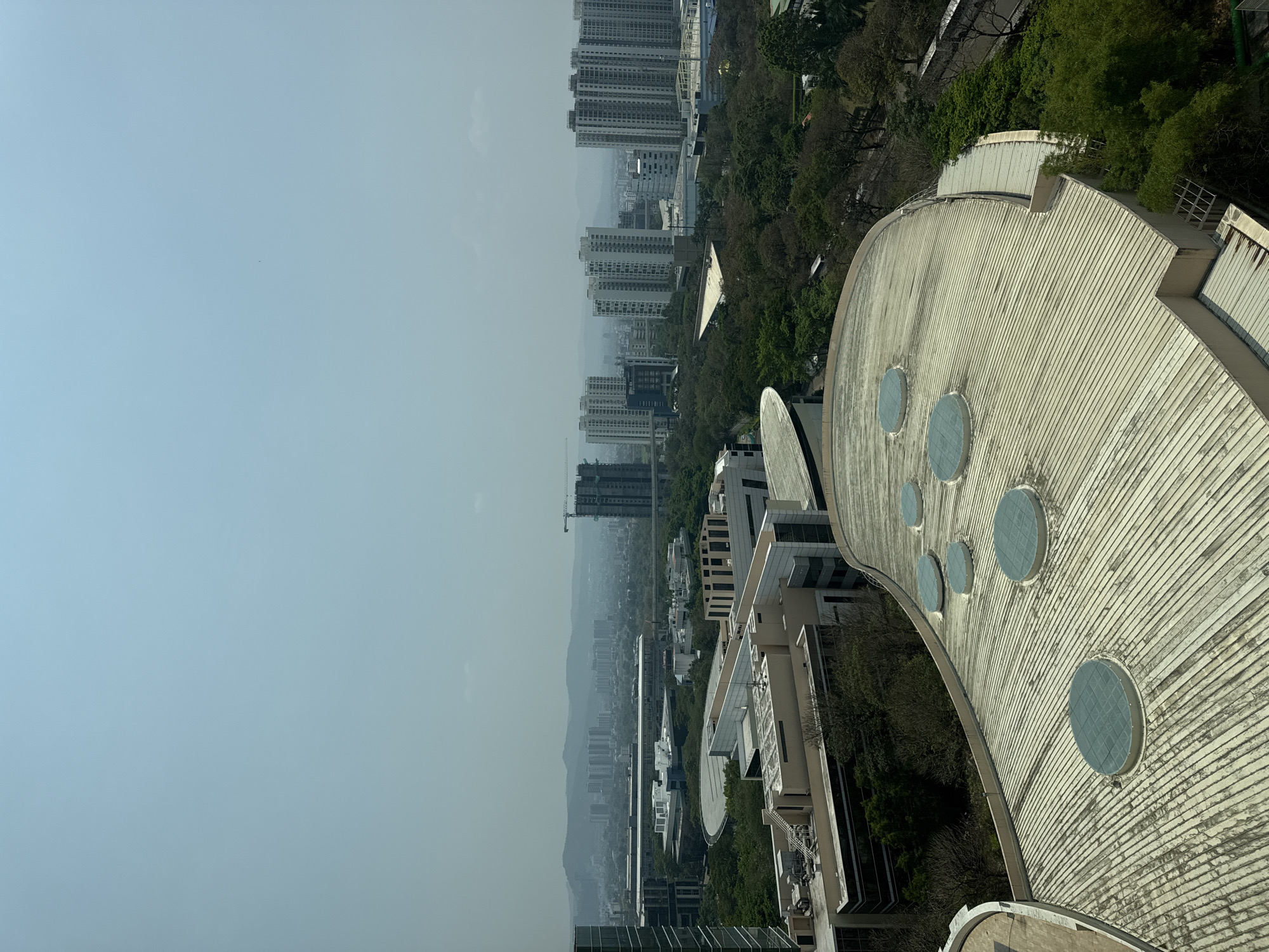 The image presents a high-angle, daytime view of an urban landscape, likely in Man, India. The foreground features a distinctive architectural element: a curved, ribbed roof with several circular skylights. To the left of the roof are modern, multi-story buildings, some adorned with solar panels on their roofs.
In the midground, a dense collection of trees separates the closer buildings from a more distant skyline. Several high-rise apartment buildings are visible, receding into a hazy atmosphere that softens the details of the landscape. The sky is a pale blue, suggesting a somewhat overcast or smoggy day. The orientation is such that the sky and distant buildings are oriented to the left, while the foreground roof occupies the right side of the frame.