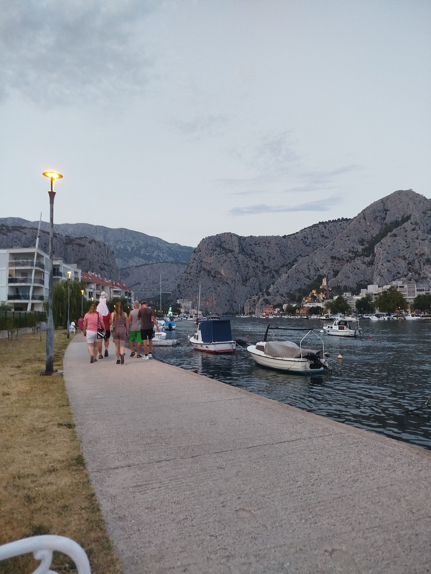 The image depicts a twilight scene along a waterfront promenade in Omiš, Croatia, with steep, rocky mountains forming a dramatic backdrop. On the left, a modern building with balconies is visible, adjacent to a grassy area. A lamppost with an illuminated yellow light stands prominently on the promenade. A group of five people, four adults and one child on someone's shoulders, are walking away from the camera along the concrete path. They are dressed in casual summer clothing.
The water is calm, reflecting the overcast sky and the lights from the town. Several boats are moored in the harbor, including small motorboats and a few larger vessels. One boat in the foreground has a canvas cover. Across the water, the town of Omiš stretches along the base of the mountains, with buildings and trees visible. A prominent fortress or castle ruin is perched high on the mountainside overlooking the town. The overall atmosphere is peaceful, with the approaching dusk casting a soft light over the scene.