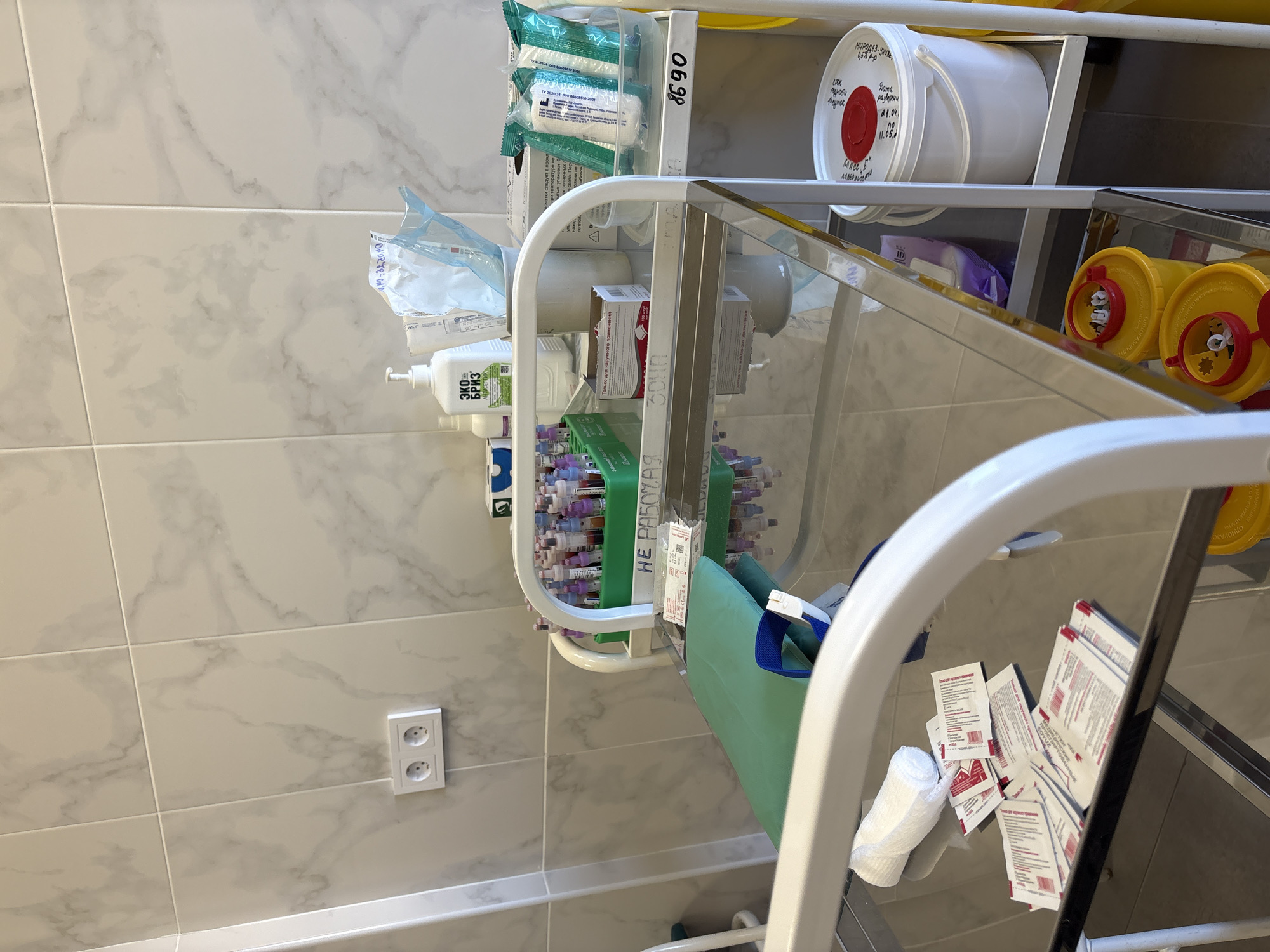 The image shows a medical cart in what appears to be a clinic or hospital room. The wall is tiled with a marble-like pattern. On the wall, there's a double electrical outlet.
The cart contains various medical supplies, including boxes of bandages, tubes, possibly blood collection tubes, and hand sanitizer. There are also containers, one of which is a white bucket with a red lid and Cyrillic text. There is also a green disposal container on the cart.
The cart has a sign reading "НЕ РАБОЧАЯ" (not working), in Russian. Some items on the cart have labeling in Russian, confirming the Russian origin of the scene. Some of the labels show Cyrillic dates, like 11.05. A hand sanitizer bottle is labelled "ЭКО БРИЗ Антисептик".
The environment is clean and sterile, suggestive of a medical setting.
Based on the use of Russian and information on the containers, the location is likely in a medical facility in Russia. Without further details, pinpointing it to Saint Petersburg is speculative.