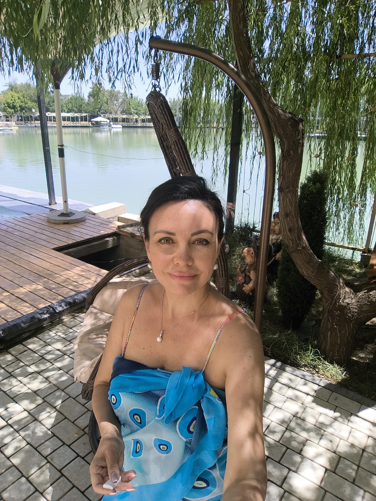 A woman with dark hair and brown eyes is taking a selfie outdoors. She is sitting in a chair under a weeping willow tree, with a body of water in the background. She is wearing a blue and white patterned sarong and a pearl necklace. The water appears to be a lake or pond, and there is a wooden dock on the left side of the image. The ground is paved with light-colored tiles. There are some garden gnomes and a small evergreen tree in the background. The overall impression is a relaxed, summery scene. The image is undated and no location is provided.