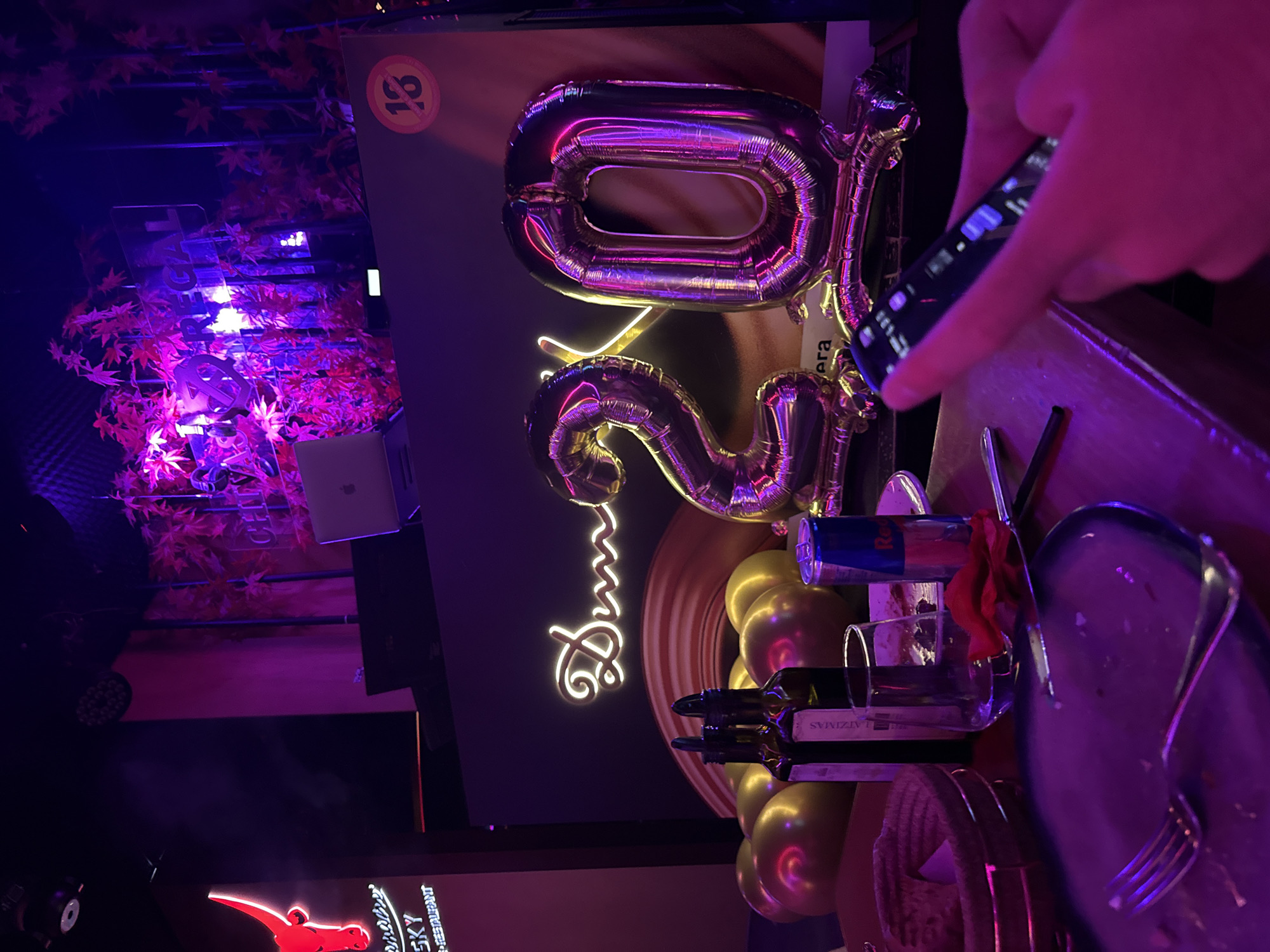 The image depicts a dimly lit indoor venue, possibly a restaurant or nightclub, with a strong purple and pink lighting scheme. In the foreground, a table is set with plates, cutlery, wine bottles, and a can of Red Bull. A hand is visible holding a smartphone. In the background, large foil balloons shaped like the numbers "2" and "0" are prominent, suggesting a celebration. A neon sign with cursive text, possibly a name or greeting, is also visible. Above the balloons, decorative elements resembling red leaves are suspended, illuminated by purple lights. To the left, a red neon bull logo with text "SKY" and "RESTAURANT" is partially visible. The overall atmosphere suggests a festive occasion, possibly a birthday. The venue appears to be "SKY" restaurant and bar located in Sofia, Bulgaria.