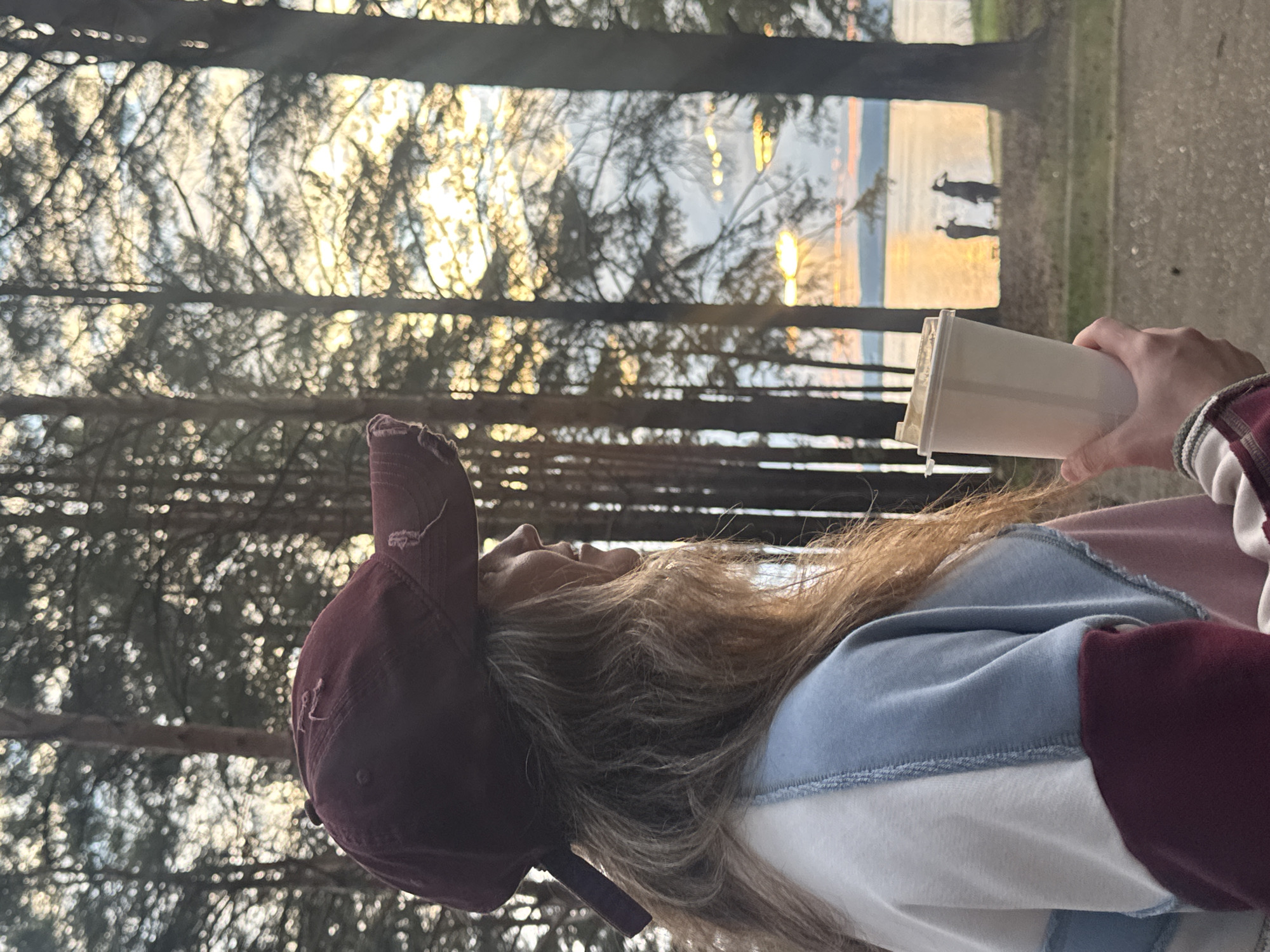 A woman wearing a maroon baseball cap and a color-blocked sweatshirt is holding a white paper cup in her right hand. She is looking towards the right, with her profile visible. The background features a forest of tall trees with the golden light of sunset filtering through them. In the distance, a body of water is visible with two silhouetted figures standing on the shore. The image appears to be taken in a park or natural area on the outskirts of the city of Chaikovsky, Russia.