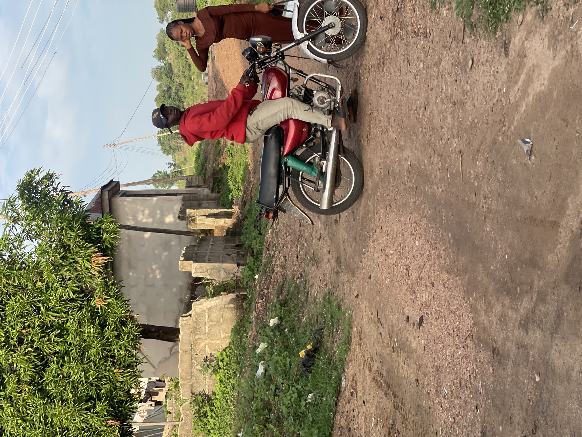 The image captures two individuals posing with a red motorcycle on an unpaved dirt path in a rural or semi-urban setting, likely in Lokoja, Nigeria. The soft, diffuse lighting suggests either early morning or late afternoon.
A man is seated on a red motorcycle, his right foot resting on the ground. He is dressed in a red hoodie, light-colored trousers, and a black baseball cap, looking directly at the camera. The motorcycle is equipped with spoked wheels, chrome rims and handlebars, a black seat, and a chrome exhaust pipe, part of which is wrapped with green tape.
Behind and slightly to the left of the motorcycle stands a woman, also gazing towards the camera. She wears a dark brown long-sleeved garment, possibly a dress, and has braids. Her right hand is relaxed near her face, and a white bag is slung over her left shoulder.
The immediate surroundings consist of a reddish-brown dirt path. To the left, concrete block structures, some appearing incomplete or aged, line the path. A large, leafy green tree dominates the far left of the frame. The ground adjacent to the path features patches of grass interspersed with some litter, including plastic bags.
The background reveals a clear, bright sky traversed by utility poles and power lines. Further in the distance, rolling hills covered with dense green foliage complete the landscape. Both individuals appear calm, seemingly posing for the photograph. No discernible text is visible on any objects or clothing.