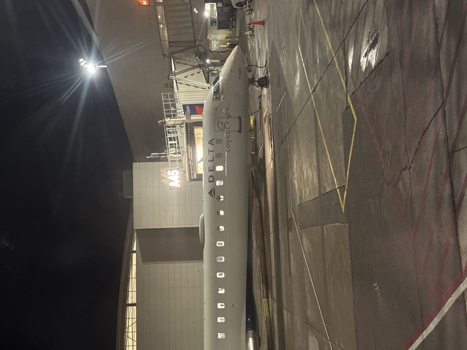 A Delta Connection airplane is docked at gate A45 at night. The plane is white with the Delta logo and "CONNECTION" displayed on its side. Exterior lights illuminate the plane and the gate area. Yellow and red lines are painted on the tarmac, which appears wet. The gate area is well-lit, contrasting with the dark sky and surrounding environment.