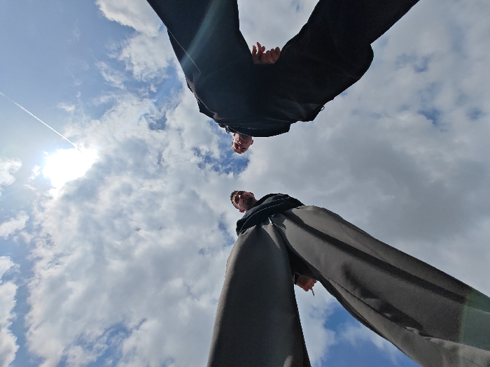 A low-angle photograph captures two individuals, presumably males, standing and looking downwards towards the camera's position against a partially cloudy sky in Shumen, Bulgaria. The sun is visible in the upper left, casting a bright glare and illuminating white cumulus clouds across a blue sky, with a faint contrail visible diagonally above the sun.
The first individual is positioned centrally at the top of the frame, viewed from the waist down, wearing dark trousers and a dark top. Their hands are partially visible near their midsection. The second individual is positioned at the bottom right, also viewed from the waist down, wearing dark trousers and a dark top. Their right hand is visible, potentially holding a small, dark, cylindrical object. Both individuals' faces are visible, looking directly downwards towards the point of the photograph. No other specific objects or distinct environmental features are discernible.