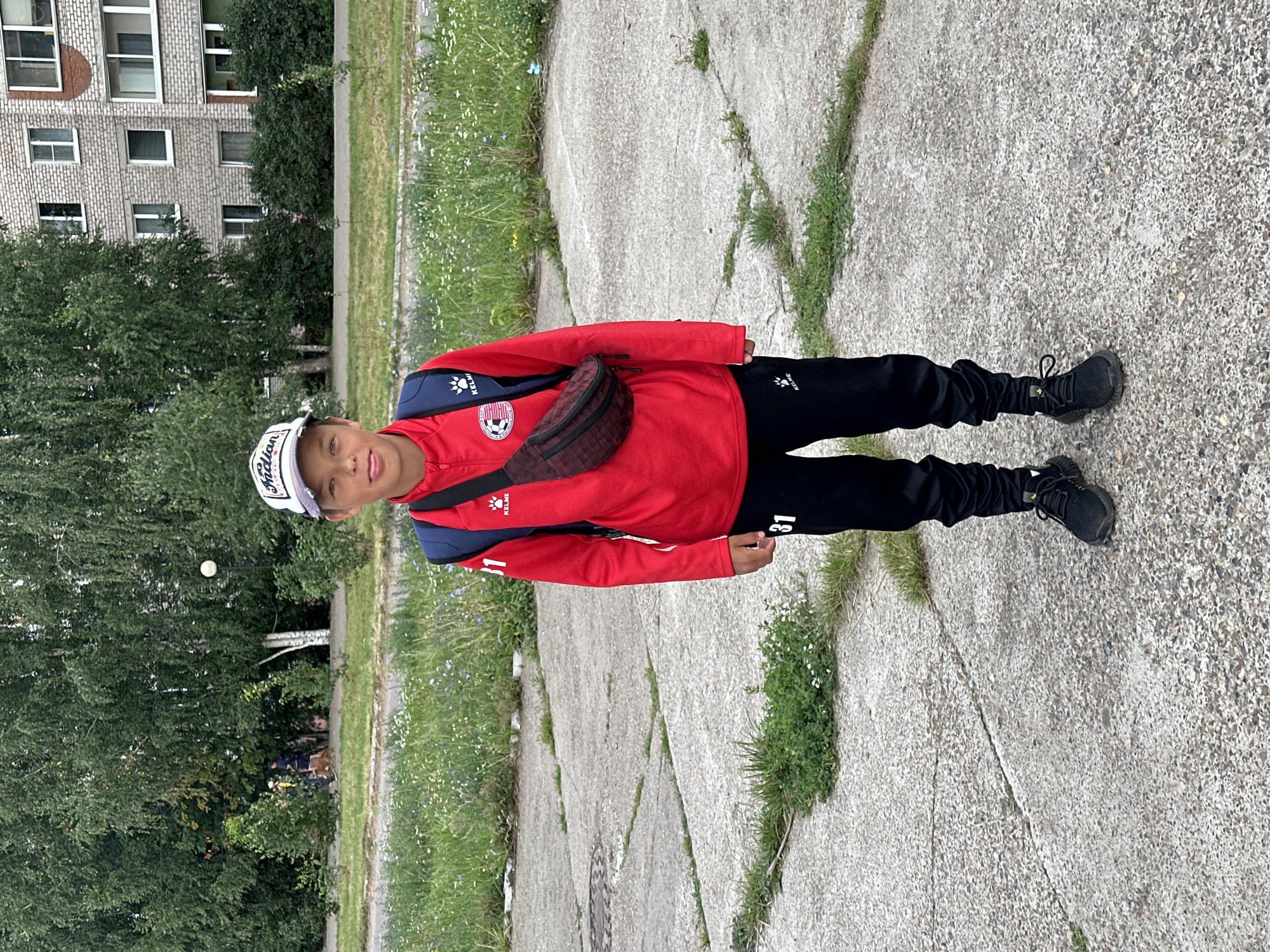 A young boy stands on a concrete paved area in front of a building and trees. He is wearing a red long-sleeved shirt with a logo on the left breast, black pants with a logo on the right thigh, and black sneakers. He has a blue backpack on his shoulders and a dark red fanny pack around his waist. He is wearing a white baseball cap with black lettering. The building in the background appears to be a multi-story apartment building, with several windows visible. Trees and green grass are also present in the background. The concrete area has cracks and some patches of grass growing through it. The sky is overcast.