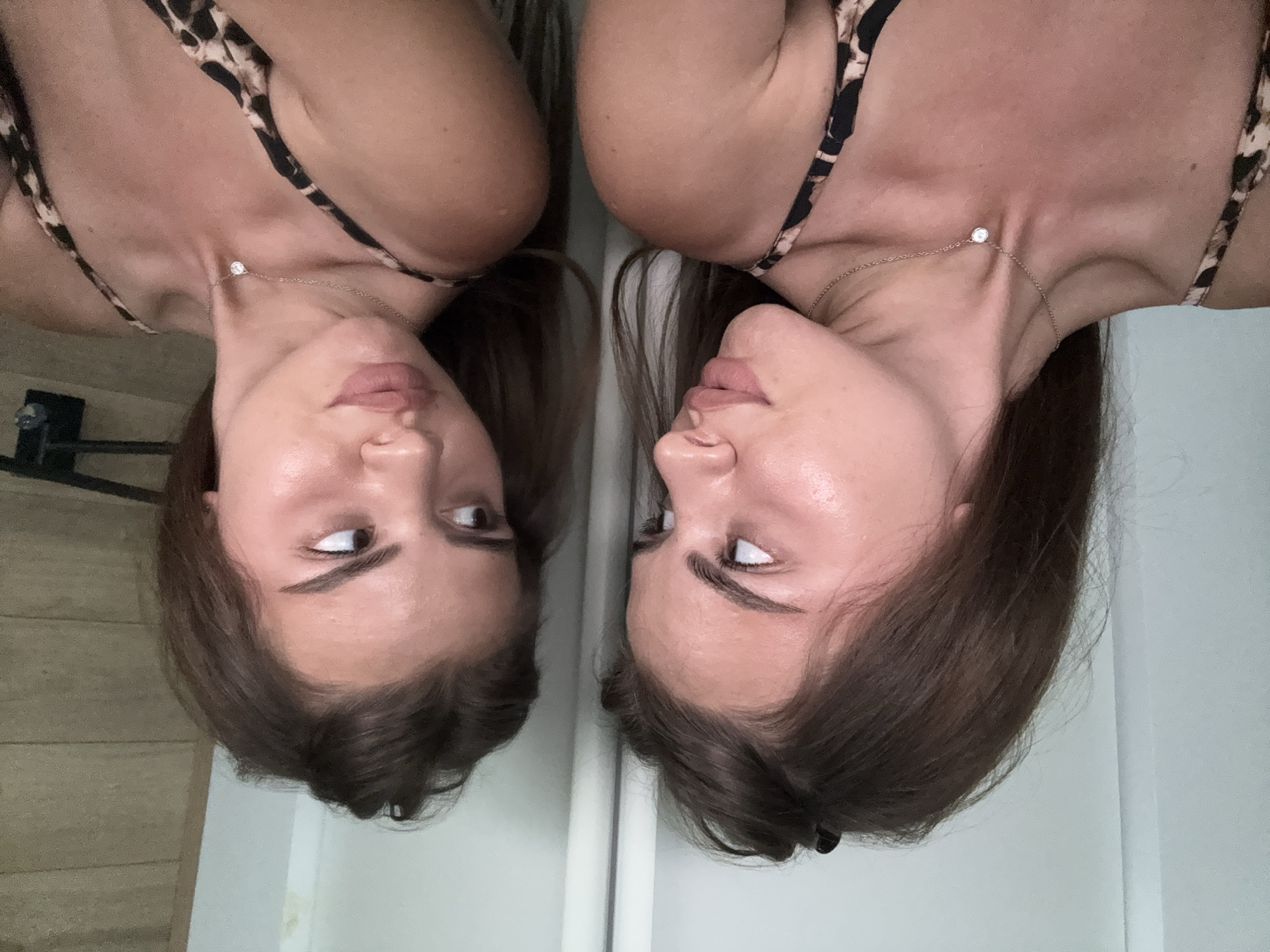 A woman is seen in front of a mirror, creating a mirrored reflection. She is wearing a top with a leopard print design and a delicate necklace with a single stone. Her hair is brown and neatly styled. The background shows a simple interior with a glimpse of a wooden panel. The location is Unknown, Unknown.