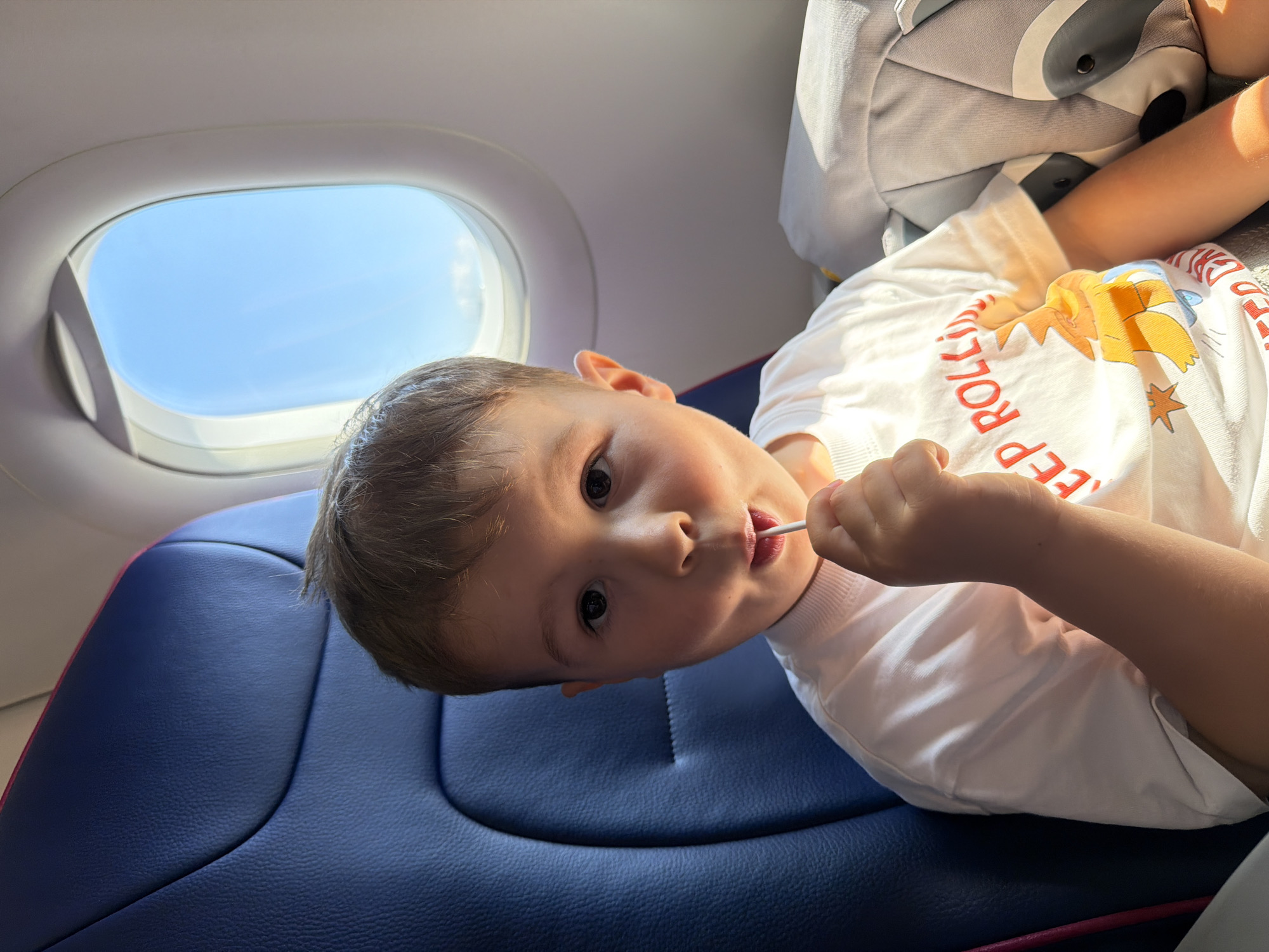 A young boy sits in an airplane seat, holding a lollipop to his mouth. He is wearing a white t-shirt with a graphic design and red lettering. To his right, a grey backpack with a raccoon design is visible. Behind him, a blue airplane seat is present, and to his left, an airplane window shows a bright blue sky with scattered clouds. The scene appears to be within the cabin of a commercial aircraft, likely en route to or from Donja Cijevna, Montenegro, given the context provided.