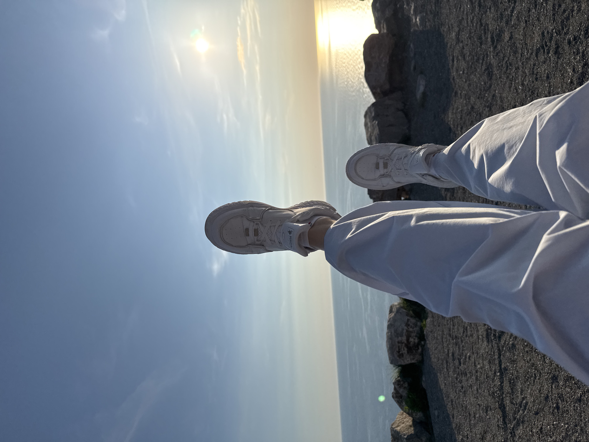 The image displays a person's lower legs and feet, clad in white loose-fitting pants and white athletic sneakers, extended horizontally across the frame. The scene is captured from a high vantage point, looking out over a vast body of calm water, likely the sea, which stretches to the horizon. The bright sun is low on the horizon, casting a strong, luminous reflection across the water, indicative of either sunrise or sunset. The sky is a clear, light blue with sparse, subtle white clouds. To the right and beneath the extended legs, dark, rugged rock formations delineate the edge of a cliff or elevated terrain. A small, indistinct logo is visible on the side of the left sneaker. The overall ambiance is peaceful and contemplative, showcasing an expansive natural vista from Kovačko Polje, Montenegro.