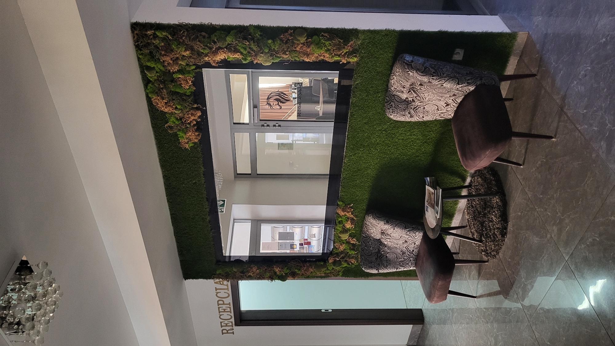 This is an indoor shot of a reception area in Prešov, Slovakia. The focal point is a large mirror mounted on a wall covered in artificial grass and moss. Two plush chairs with patterned backs and dark brown seats are positioned in front of the mirror. A small, dark circular table with a few magazines sits between the chairs. To the left, a door with the word "RECEPCIA" above it is visible, indicating "Reception" in Slovak. The floor is tiled with a grey, marbled pattern. A modern chandelier hangs from the ceiling. The mirror reflects an interior space with glass doors and a sign displaying a horse logo.