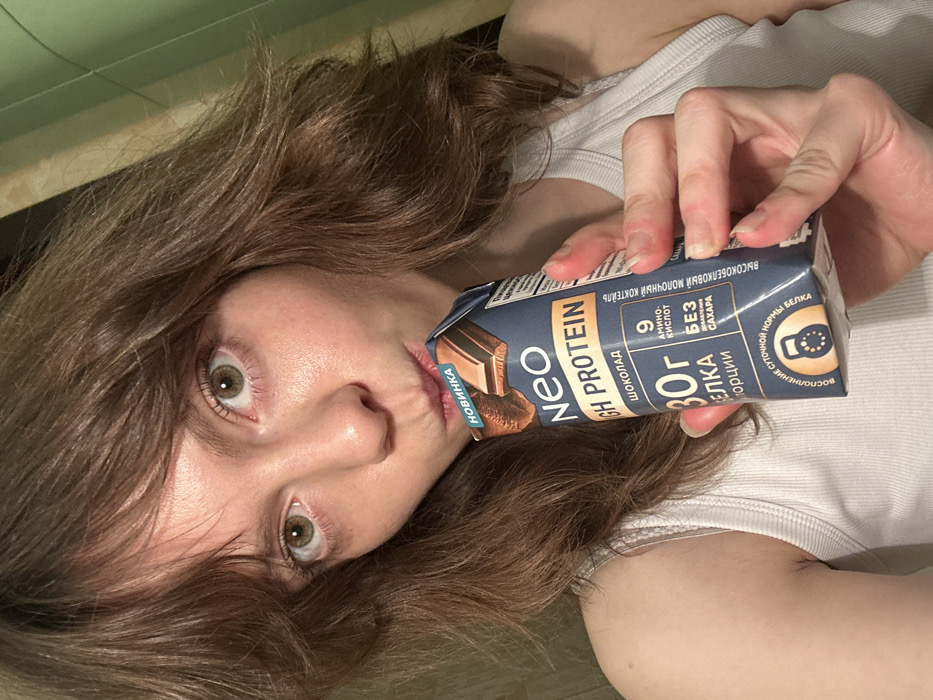 A close-up, chest-up selfie depicts a young adult female indoors, actively consuming a protein drink. The woman has shoulder-length wavy brown hair with bangs and light-colored eyes, directly gazing at the camera. She is wearing a white ribbed tank top. Her lips are puckered around the opening of a rectangular, dark blue "neo HIGH PROTEIN" chocolate-flavored drink carton, which she holds in her right hand. The carton features Russian text, including "НОВИНКА" (New Product), "ШОКОЛАД" (Chocolate), "30Г БЕЛКА" (30g Protein), "9 АМИНОКИСЛОТ" (9 Amino Acids), and "БЕЗ САХАРА" (Sugar-Free). The background consists of indistinct indoor surfaces: a light brown or beige vertical surface on the left and a darker, possibly green or dark blue wall on the right. No specific landmarks or architectural features are visible to precisely identify a location within the city of Moscow, Russia; however, the product labeling is in Russian.