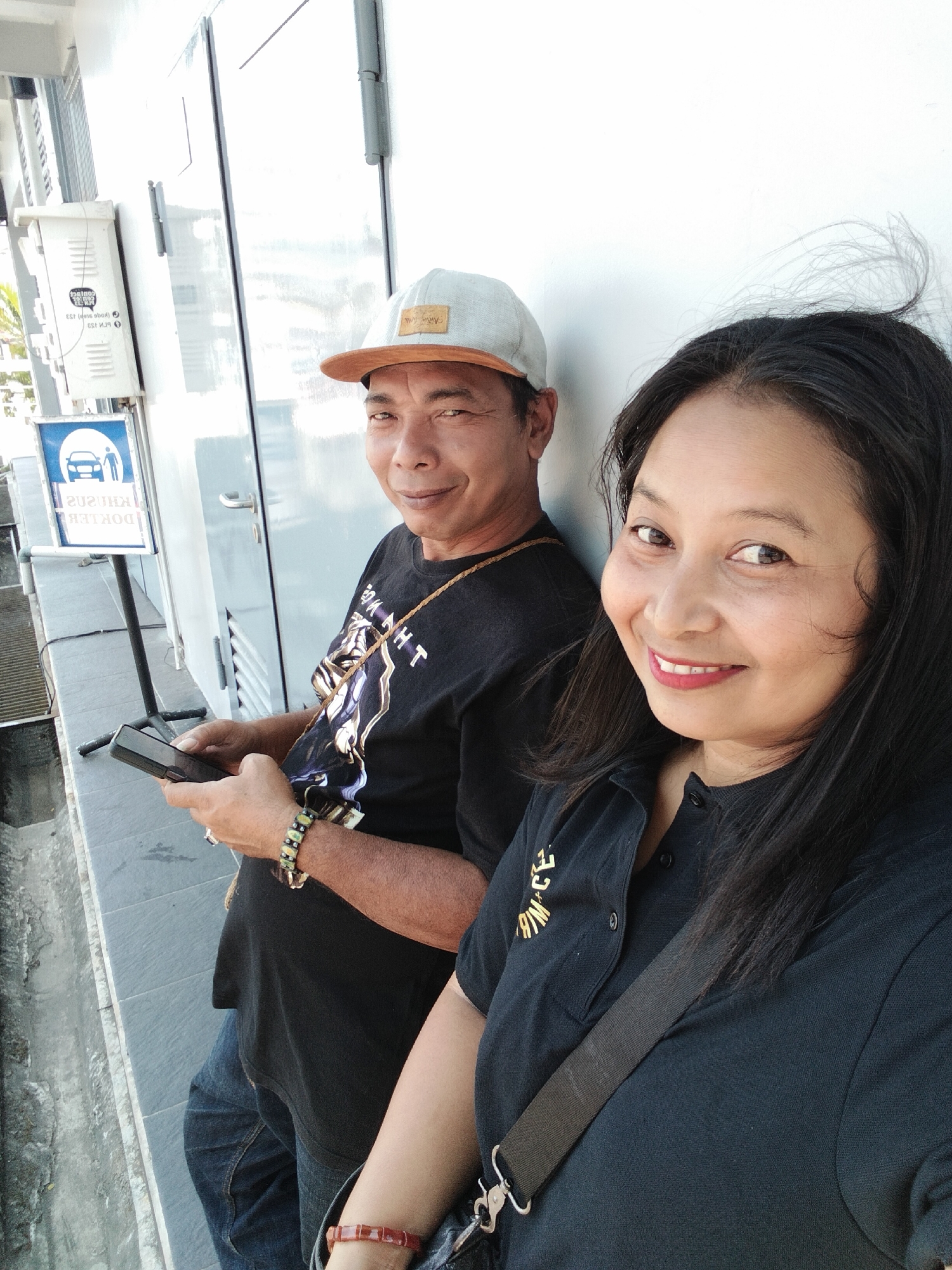 This is a selfie of a man and a woman in the city of Magelang, Indonesia. The man, wearing a black t-shirt and a cap, is holding a smartphone. The woman, in a black polo shirt, is smiling at the camera. They are standing next to a white wall, possibly outside a building. A blue sign with a car and pedestrian icon is visible in the background, indicating a parking or drop-off zone. The overall impression is a casual, everyday moment captured by the couple.