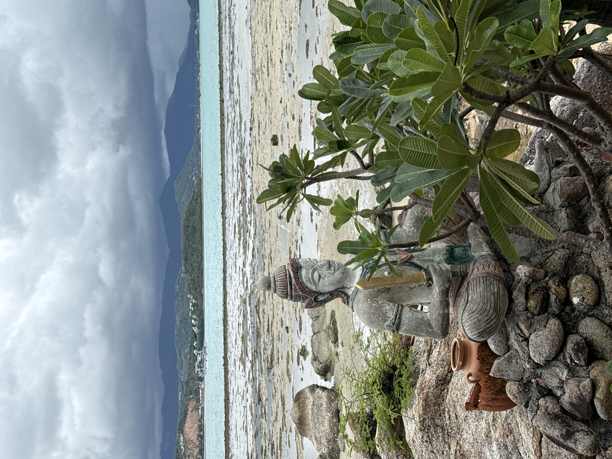 A seated statue of a figure with an ornate headdress and traditional clothing is positioned on a rocky outcrop overlooking a shallow, tropical body of water. The statue is light-colored with painted details on its headdress and clothing. To the left of the statue, resting on the rocks, is a rustic, terracotta-colored vase with two handles. In front of the statue, a leafy green plant with broad leaves grows from the rocky terrain. The background features a wide expanse of shallow water, dotted with patches of exposed sand or rocks, indicative of a low tide. Beyond the water, a distant coastline is visible, lined with green hills and mountains under a cloudy, overcast sky. Buildings and what appear to be structures are scattered along the distant shoreline, suggesting a populated area. The overall scene depicts a tranquil, natural setting with a cultural element. The location is identified as Ko Samui District, Thailand.
