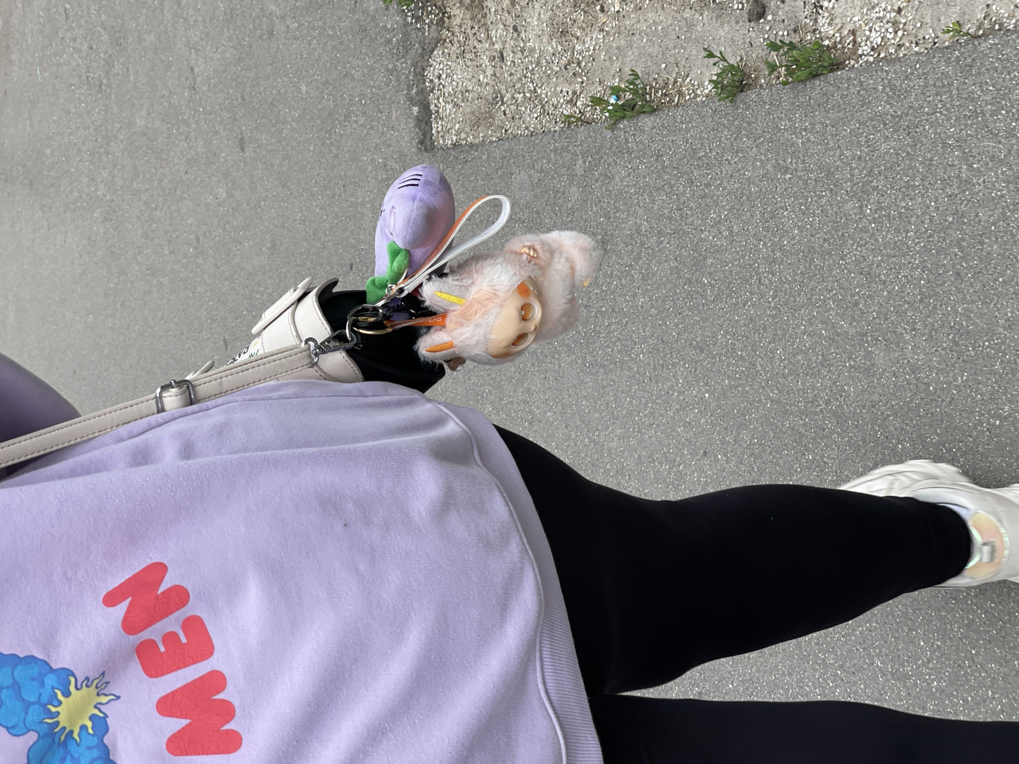 A person wearing black leggings and a light purple sweatshirt with "MEN" written in red is walking on a paved path. The sweatshirt has a blue and yellow graphic on the left shoulder. The person is carrying a white purse with a silver buckle. Attached to the purse are two keychains: a small, fluffy, light orange creature with big eyes and a purple plush toy resembling a vegetable with a green stem. The paved path is bordered by a strip of dirt and small green plants. The scene appears to be outdoors, possibly in a park or along a street.