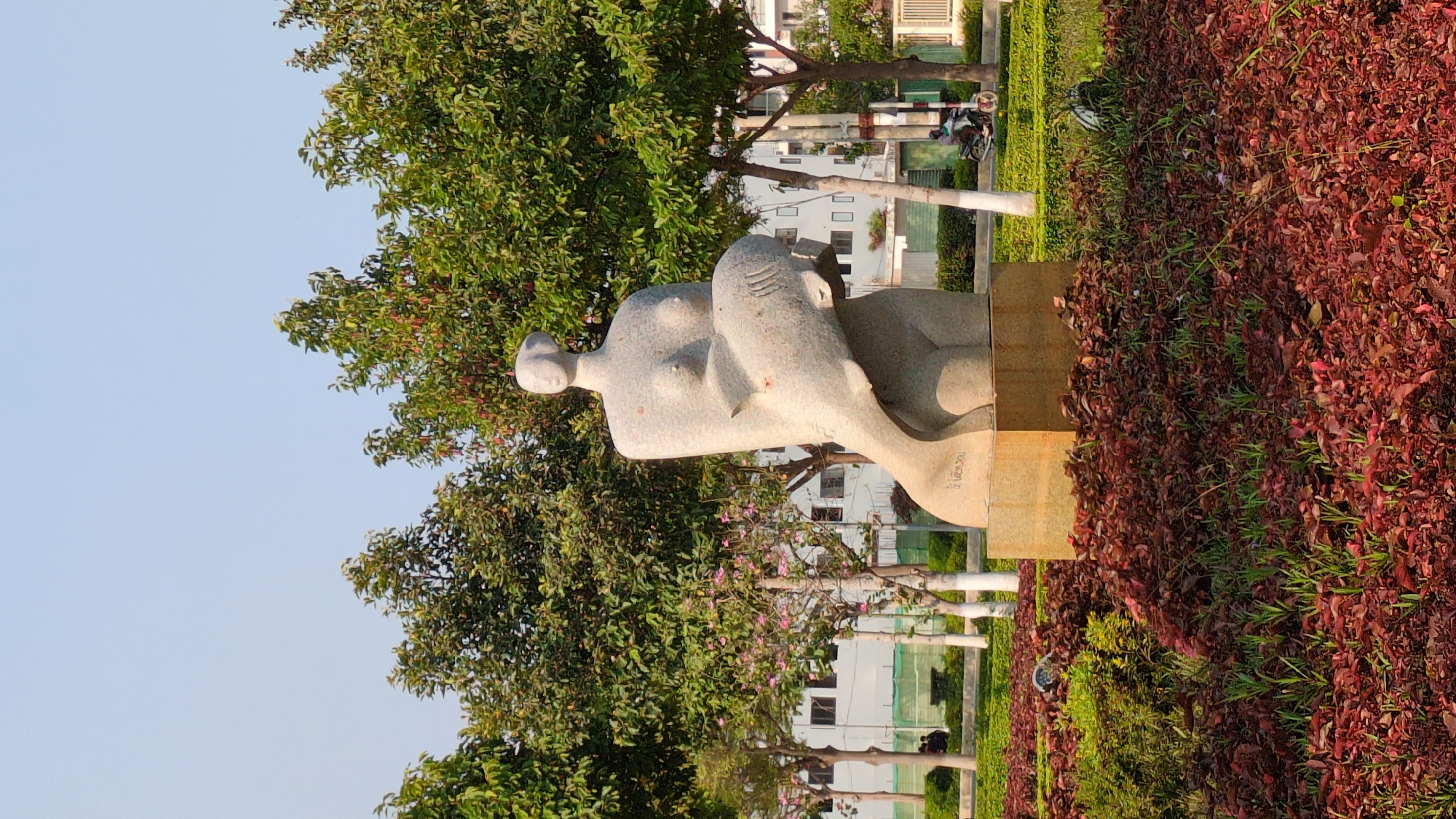 The image features a marble sculpture of a reclining woman with a curved, smooth form placed on a stone pedestal, situated in a park setting. The sculpture sits amidst lush greenery, including trees and what appears to be a dense bed of red-leaved ground cover. In the background, there are white buildings with windows and a glimpse of what may be a street with utility poles and possibly a motorbike. The sky is light blue, suggesting either early morning or late afternoon light. The location is indicated as being in the city of Phan Rang – Tháp Chàm, Vietnam.
