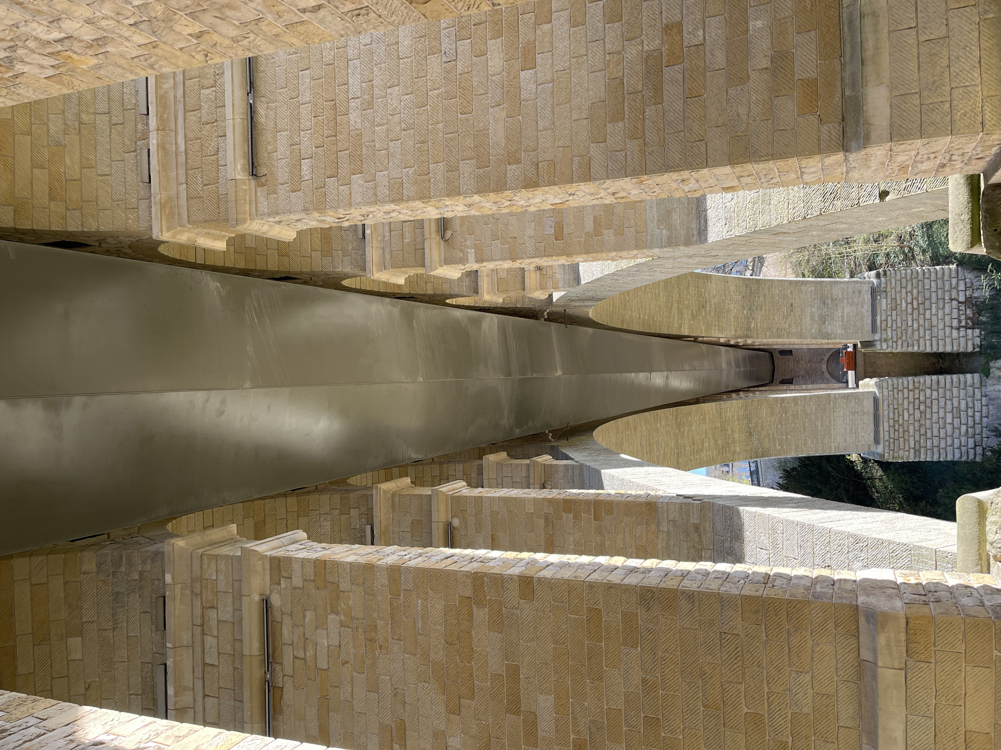View from underneath the Pont Adolphe in Luxembourg City, Luxembourg. The bridge is constructed of light-colored stone and features multiple arched supports. A steel plate spans the bridge's base. Vegetation and other bridge structures can be seen below the bridge.