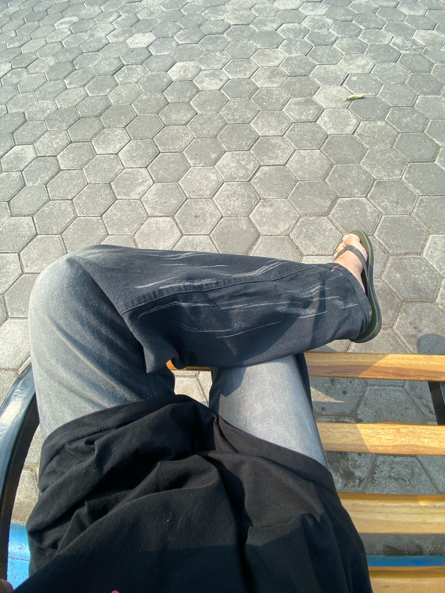 The image shows a person's legs sitting on a wooden bench. The person is wearing grey jeans and dark pants with white lines on them, and green flip-flops. The ground is paved with hexagonal grey tiles. The scene appears to be outdoors, possibly in a public space like a park or walkway, in Sidoarjo, Indonesia, judging by the hexagonal tile pattern often found in such areas. Sunlight casts shadows on the ground and bench.