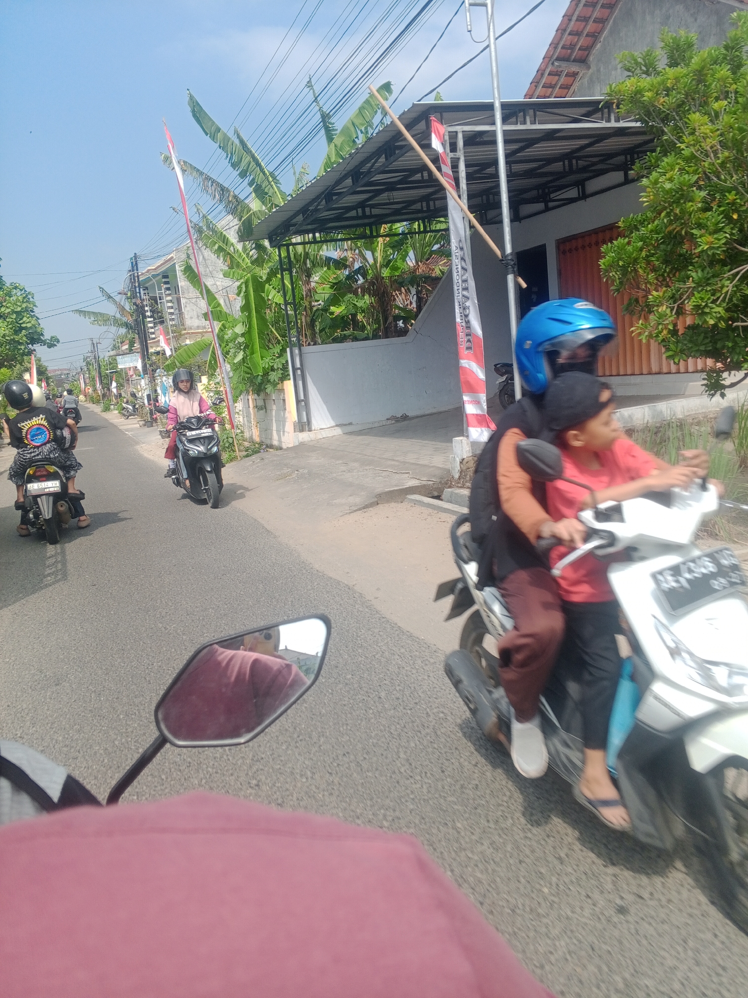 In Ronowijayan, Indonesia, on a bright, clear day, the scene captures a residential street with various motorcycles in motion. The foreground features the perspective of a rider, with a pink garment and a side mirror visible. To the right, a white scooter is being ridden by a person in an orange jacket and brown pants, with a child wearing a red shirt and black cap as a passenger. The child is looking to the right. Behind them, another motorcycle carries two individuals, one wearing a black shirt with a graphic design and a black helmet, and the other in a pink and white outfit with a helmet. Further down the street, another motorcycle is visible, with a rider wearing a black helmet. The surroundings include houses with tiled roofs, one with an orange shuttered garage door, and lush green trees and banana plants. Indonesian flags are displayed along the street, attached to poles. The road surface is asphalt, and a sidewalk is present. Electrical wires are strung overhead.