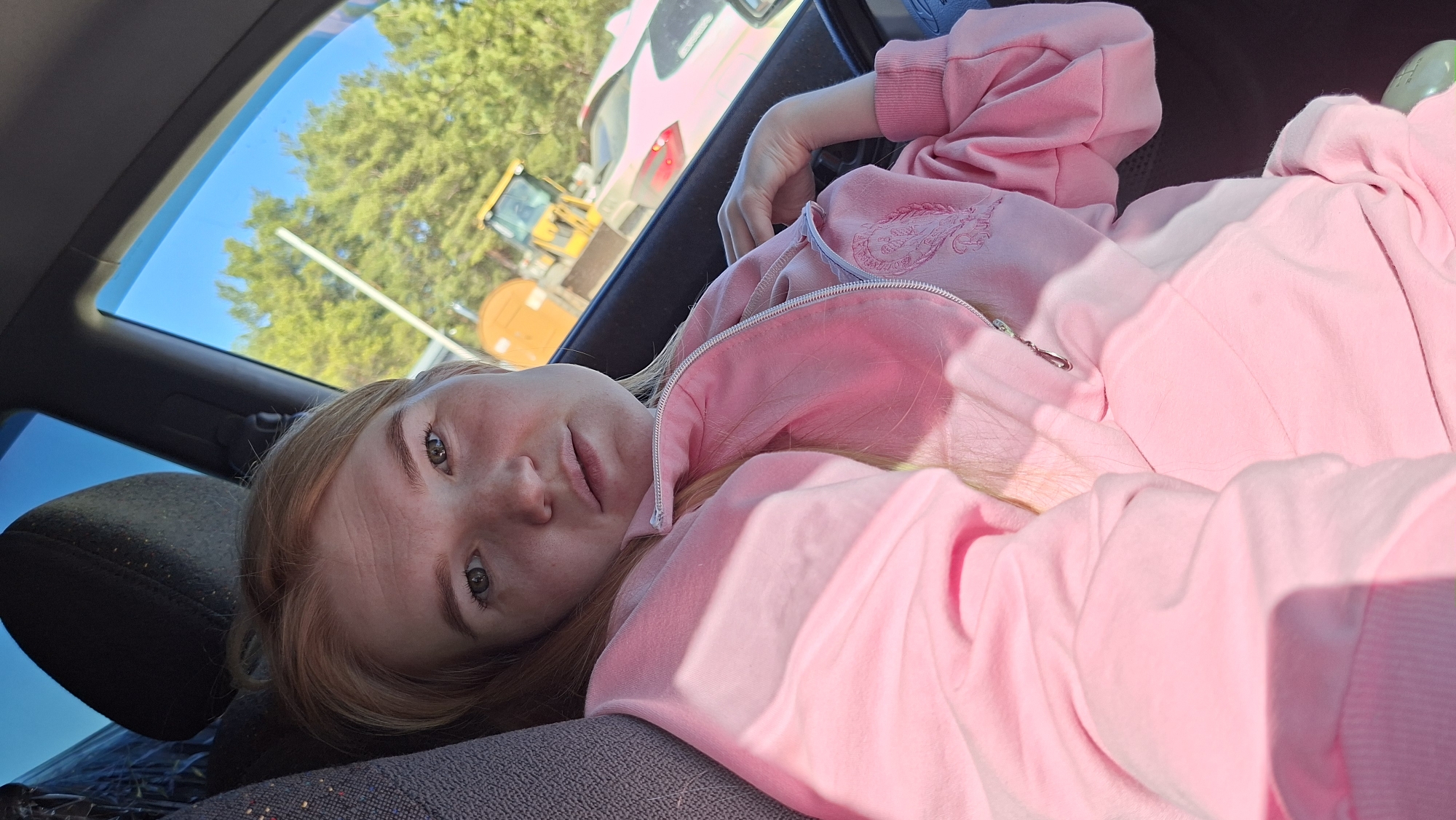 A young woman is lying in a car, wearing a pink tracksuit. Outside the car window, a construction site is visible, featuring a yellow construction vehicle. The location is Chaikovsky, Russia.
