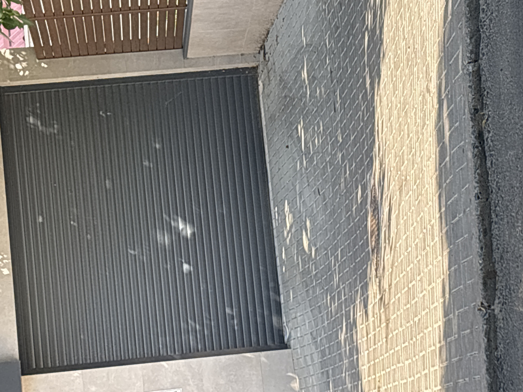 The image shows a closed gray roller shutter door, likely for a garage or commercial establishment. The door is installed on a building with light-colored tiled walls. In front of the door is a paved driveway or walkway made of light gray rectangular bricks laid in a pattern. Sunlight casts dappled shadows on the paved surface, indicating an outdoor setting. To the right of the roller shutter, there is a section of a brown wooden slatted fence. The scene appears to be a typical urban or suburban environment.