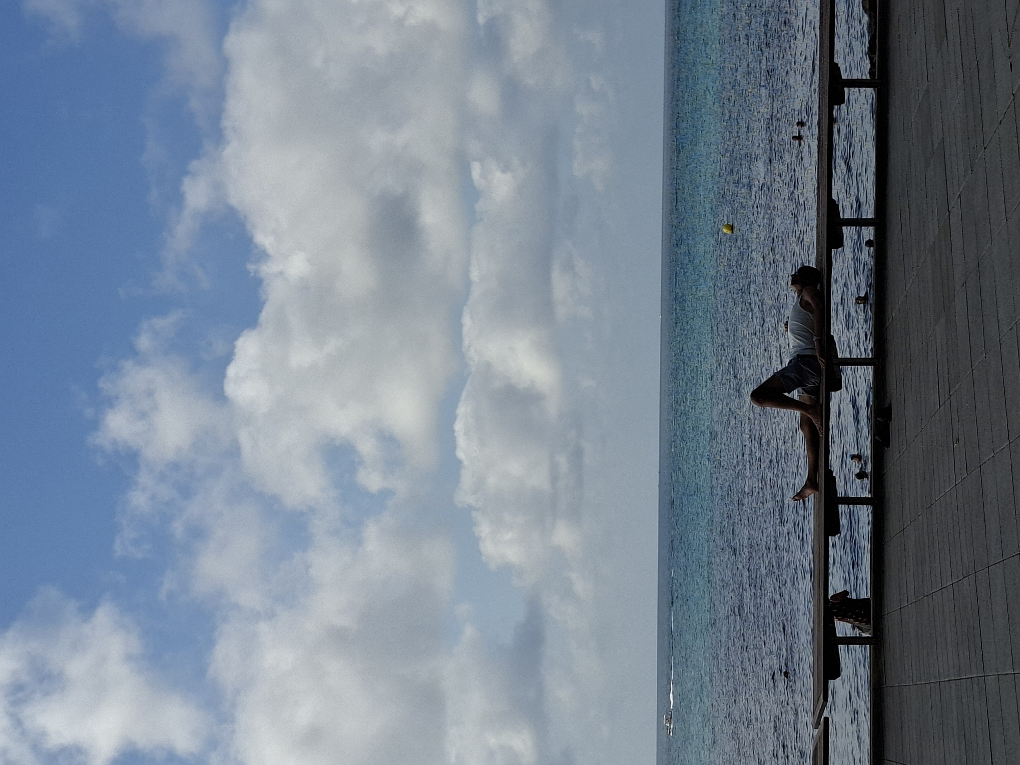 The image depicts a person lying on a wooden bench facing upwards, with their eyes closed and head tilted back. They are wearing a white tank top and dark shorts. The bench is positioned along a promenade overlooking a calm, blue body of water, likely the Mediterranean Sea. In the distance, a small boat is visible on the water, and a yellow buoy floats nearby. The sky above is a clear blue with scattered white clouds. The foreground shows a grey tiled walkway. The scene appears to be a tranquil moment of relaxation in a coastal setting, characteristic of locations like the promenade along the coast of Torrevieja, Spain.