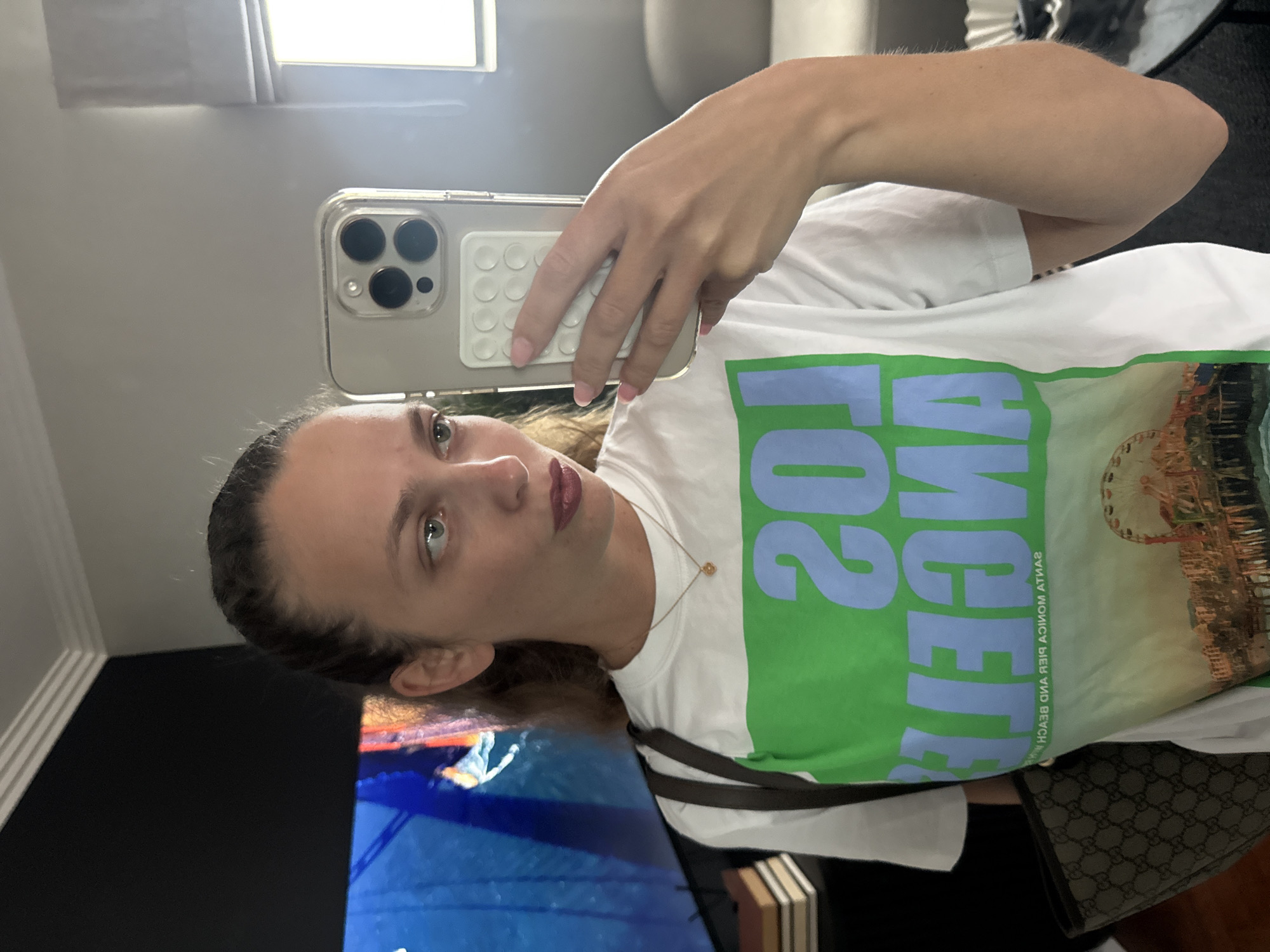 A woman is taking a selfie in a mirror. She is holding a phone with a clear case and a white grip on the back. She is wearing a white t-shirt with a green and blue Los Angeles design. She has dark hair and fair skin. There is a window in the background with light coming through it. There is also a television screen reflecting blue light. A purse with a brown strap is visible. The location cannot be determined based on the image.