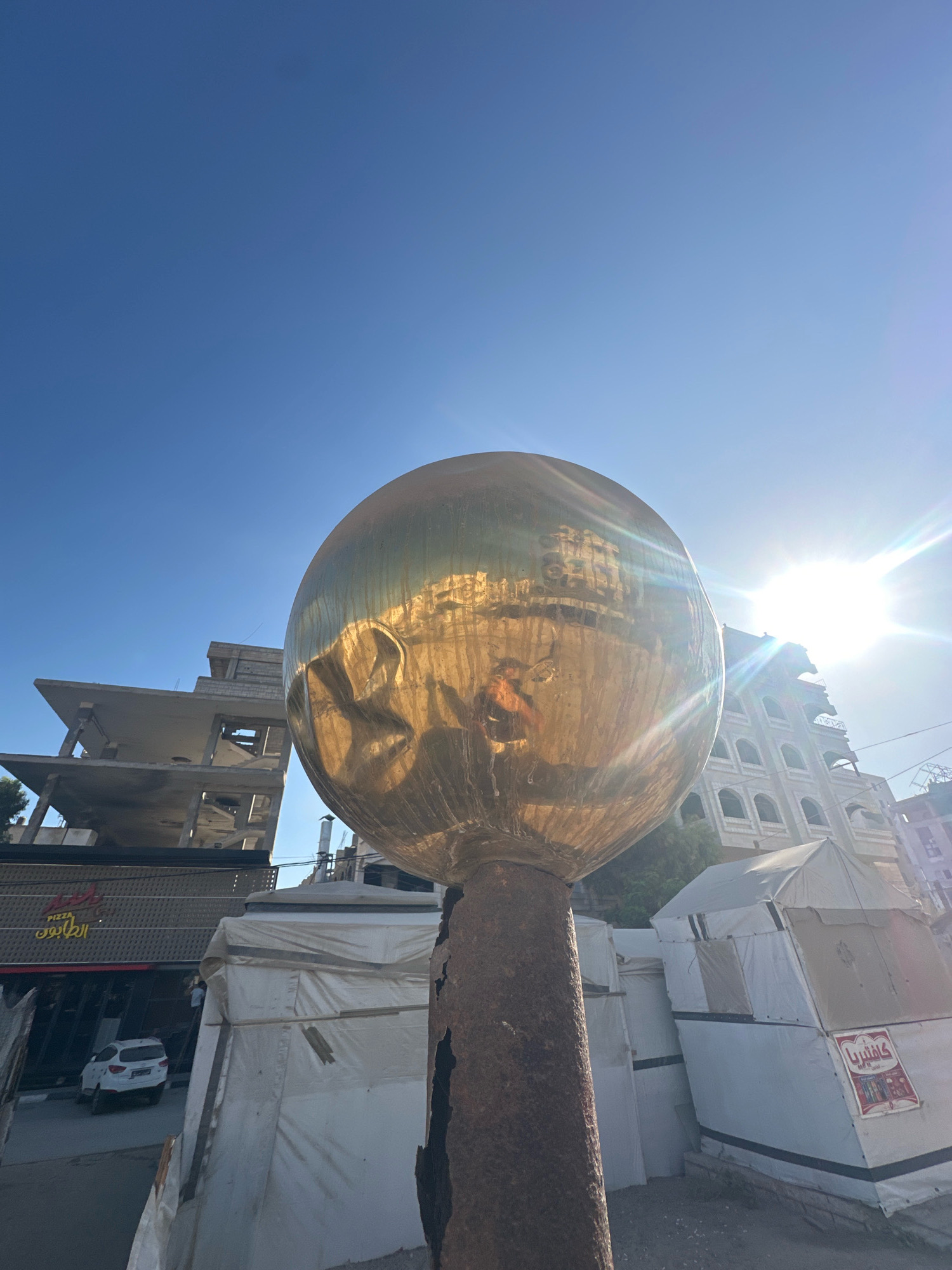 In Gaza City, Palestinian Territories, on a bright, sunny day, a close-up, low-angle shot captures a large, spherical, golden sculpture dominating the foreground. The sculpture, possibly made of polished metal, is dented and weathered, supported by a thick, rusty, and corroded cylindrical base. Reflections of buildings and the sky are visible on its curved surface.
In the background, a partially constructed building with exposed concrete floors and columns stands to the left. To the right of the sculpture, a multi-story building with arched windows and a pale facade is visible. In front of this building, several white tent-like structures are erected. One of these tents displays a poster with Arabic writing and images. A white car is parked on a street below the partially constructed building, and signage with Arabic text, including the word "PIZZA", is visible on a building facade. The sky is a clear, vibrant blue, with the sun shining intensely, creating lens flare effects. The scene appears to be an outdoor urban environment, potentially a public space or street.