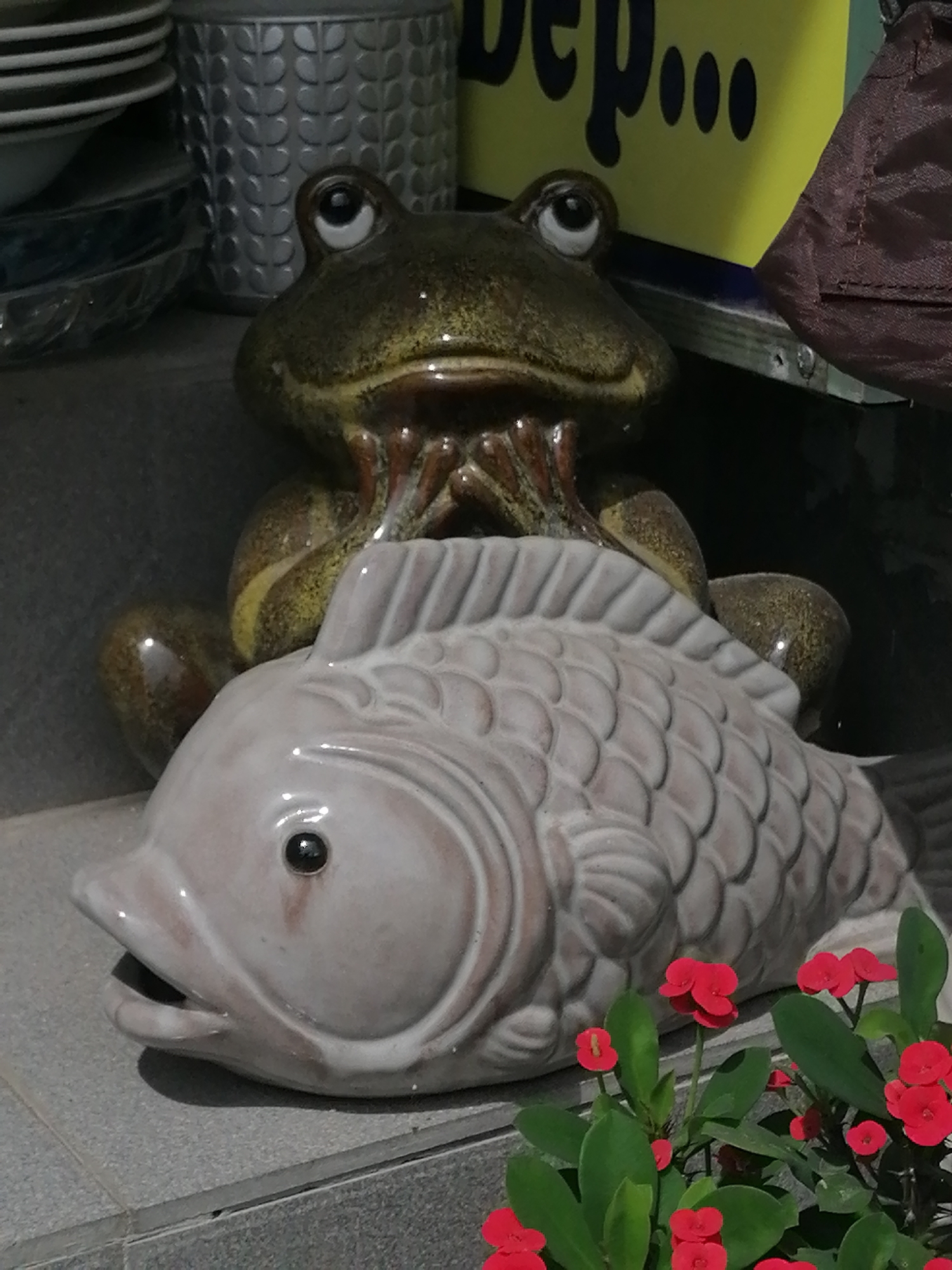 A ceramic statue of a frog is perched atop a ceramic statue of a fish. The frog is a mottled green and brown, with large, expressive eyes. Its arms are clasped in front of its chest, resting on the fish's head. The fish is a pale gray, with an open mouth and a textured body designed to resemble scales. The statues are placed on a gray tiled surface. In the foreground, a cluster of vibrant red flowers with green leaves grows in front of the fish statue. In the background, a tall, ribbed silver cylindrical object, possibly a planter or container, is visible. To the right of this object, a portion of a yellow sign with black lettering that appears to read "dep..." is visible, partially obscured by a draped dark red fabric. The scene suggests an outdoor setting, possibly a garden or a shop display, given the combination of decorative items and plant life. The presence of the "dep..." sign could indicate a location in Vietnam, perhaps a tourist area or a shop.