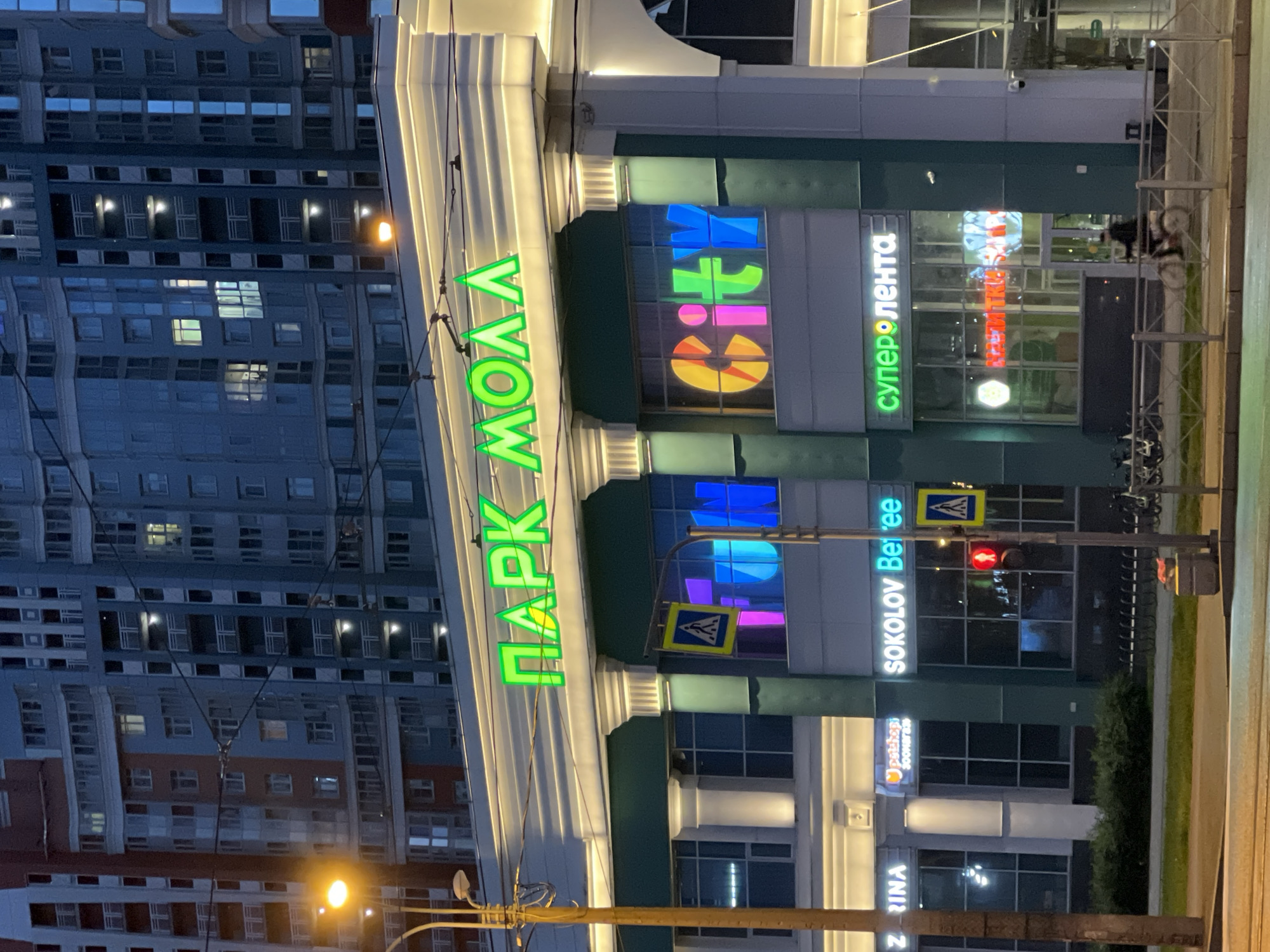This media file shows the exterior of a modern building at dusk. The building appears to be a shopping mall or commercial complex, featuring large glass windows and illuminated signage. Dominating the facade is a bright green neon sign that reads "ПАРК МОЛЛ" (Park Mall) vertically. To the right of this, another sign displays colorful, illuminated letters forming the word "CITY". Further to the right, a green neon sign reads "суперлента" (superlenta). Below these, white text on a dark background spells out "SOKOLOV Beree".
The surrounding environment suggests an urban setting with a tall residential building visible in the background, reflecting the lights of the commercial structure. A street and sidewalk are present in the foreground, with a traffic light showing a red signal. There are bicycles parked near a railing on the right side of the frame.
The time of day is clearly evening or dusk, as indicated by the dim ambient light and the illuminated signs. The weather appears to be clear, with no visible signs of precipitation. The scene evokes a sense of urban activity and commerce, with the bright lights of the mall contrasting with the darkening sky. The overall impression is one of a bustling commercial area.