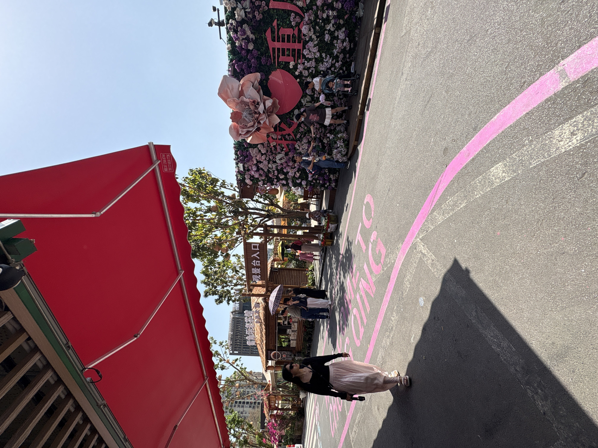 Here is a detailed description of the image:
**Scene:**
The image shows an outdoor event or market setting, possibly a festival or fair in the Yuzhong District, China. The viewpoint is from the street level.
**Environment and Objects:**
* **Road:** The road surface is asphalt, marked with large pink painted circles containing Chinese text.
* **Awning:** A large red awning is visible on the left side.
* **Floral Wall:** A large floral backdrop with an oversized flower is located at the back.
* **Booths/Stalls:** There are wooden stalls or kiosks along the road, possibly selling food or merchandise. Some have signs in Chinese.
* **Trees:** Trees with green leaves are interspersed among the stalls.
* **Buildings:** Modern buildings can be seen in the background, indicative of an urban setting.
**People:**
* **Pedestrians:** Several people are walking along the road, including a woman wearing a dark top and a light pink skirt.
* **Groups:** There are groups of people congregating near the stalls.
* **Families:** Families are pictured taking photos near the floral wall.
**Actions and Interactions:**
* **Walking:** People are strolling through the market.
* **Browsing:** People are looking at the items for sale in the stalls.
* **Taking pictures:** People are posing and taking pictures in front of the floral wall.
