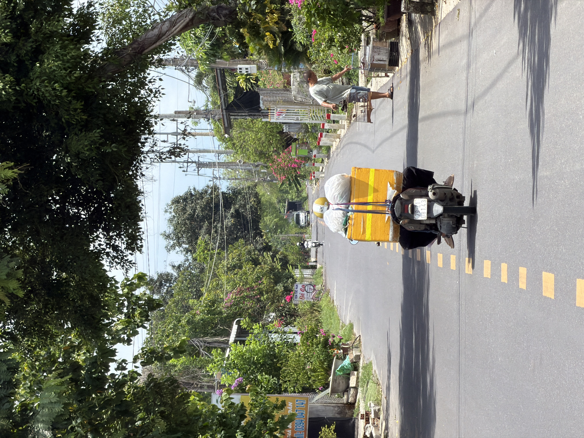 In Diên Thọ, Vietnam, a person is riding a motorbike down a paved road, carrying a large yellow box laden with white bags on the back. The rider, with their legs draped over the sides of the motorbike, is partially visible. The road is lined with trees casting shadows, and on the right side, a man in a light-colored shirt and shorts is tending to several bird cages. Buildings and more vegetation are visible along the sides of the road, with power lines overhead. The sky is a bright, hazy blue.