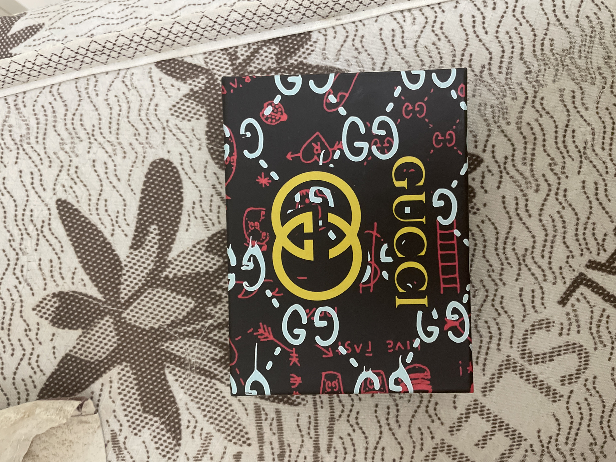 An overhead, slightly angled shot shows a dark-colored box, likely packaging, resting on a light-colored, patterned fabric surface. The box is predominantly black, featuring a vibrant, graffiti-style design. A large, yellow interlocking "GG" logo is centrally placed, with the brand name "GUCCI" written vertically in yellow capital letters on the right side of the box.
The black background of the box is embellished with numerous smaller, playful graphics in light blue and red. These include smaller interlocking "GG" logos, hearts, arrows, ladders, crowns, and various abstract doodle-like symbols. Partially visible text, such as "LIVE FAS" on the lower left, suggests a "live fast" motif.
The fabric surface beneath the box, likely a bedspread or upholstery in an indoor setting in Ihtiman, Bulgaria, has a cream or light beige base. It displays a repeating pattern of larger, abstract shapes, possibly stylized leaves or petals, rendered in a textured brown stippling. These larger forms are interconnected by wavy, dashed lines in a similar brown tone. The fabric itself appears to have a subtle woven or quilted texture. The lighting is soft and even, typical of an indoor environment.