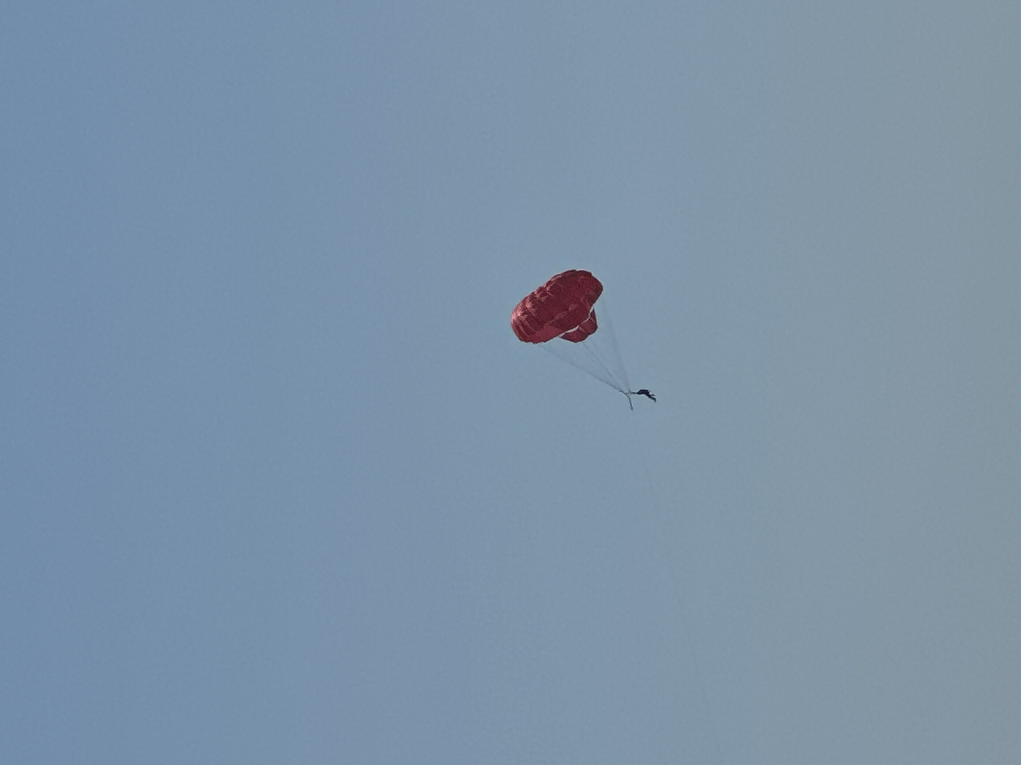 A person is parasailing high in the sky. The parachute is red and appears to be a double parachute design. The person is a dark silhouette against the bright sky, suspended by lines from the parachute. The sky is a clear, light blue, with no clouds visible. The image appears to be taken from a distance, possibly from a boat or beach. The location is Bečići, Montenegro, a popular tourist destination known for its beaches.