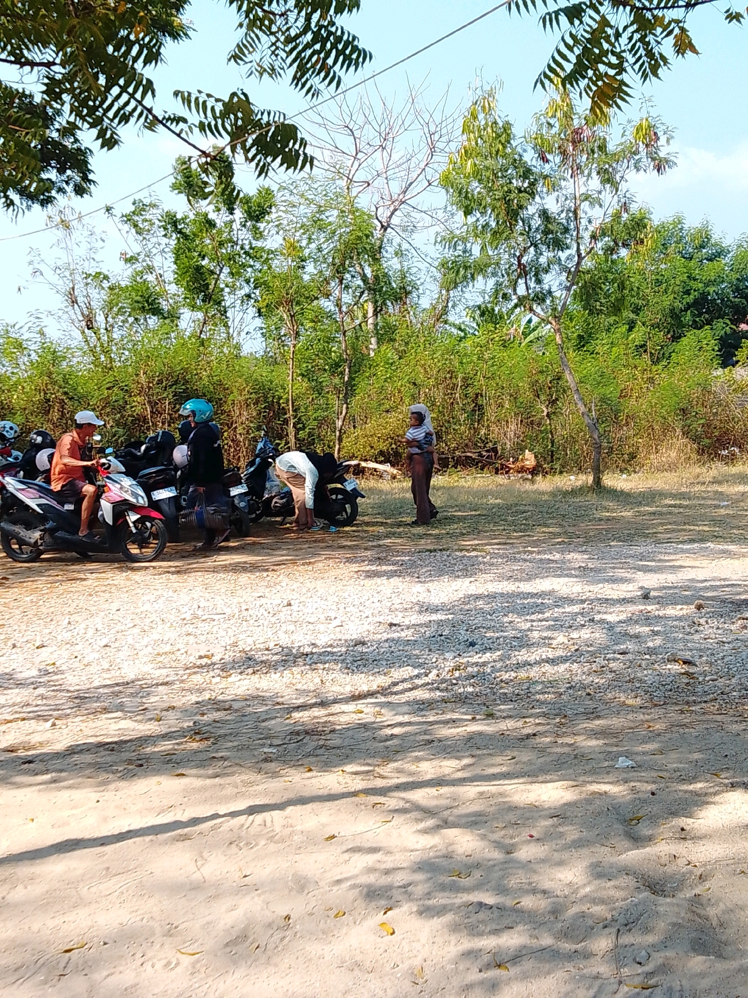 In Cikal, Indonesia, a group of people are gathered near a line of parked scooters under a clear sky. Three individuals are the main focus. To the left, a man wearing a white cap and an orange shirt sits on a red and white scooter. Beside him, another person, possibly a woman, wears a blue helmet and a dark jacket, standing near a black scooter. Further to the right, a third person, dressed in light-colored clothing, is bent over, examining something near the parked scooters. The environment is characterized by a gravelly ground and a dense green foliage in the background, with a few trees scattered throughout. The sunlight casts shadows on the ground, indicating it is daytime.