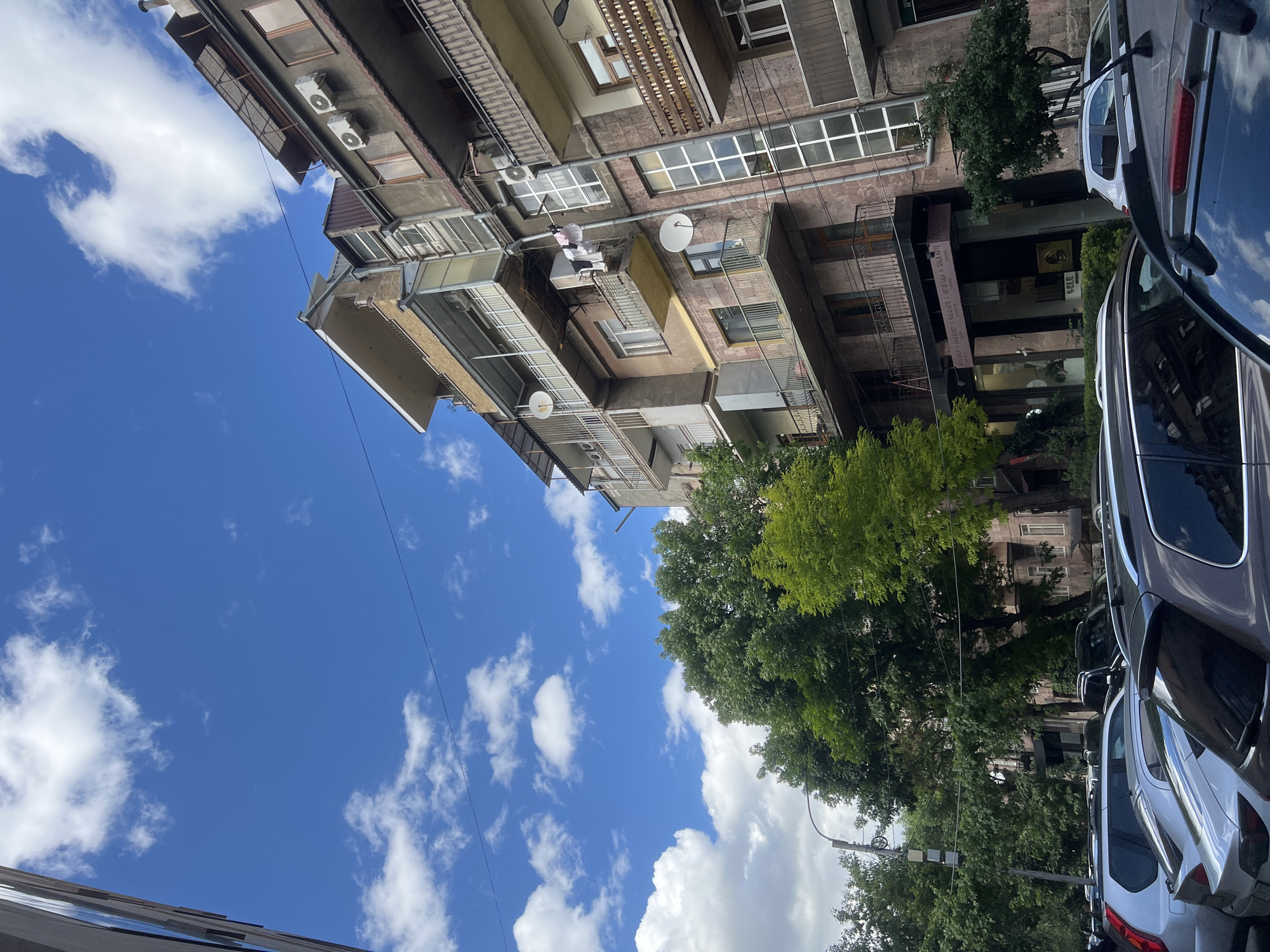 The image depicts a building in Yerevan, Armenia, under a bright blue sky with scattered white clouds. The building has multiple balconies with varying designs, some featuring satellite dishes and air conditioning units. Trees obscure parts of the building. Parked cars line the street, visible on the right side of the frame. The building has a banner that reads "ART GALLERY GALA ARTE."