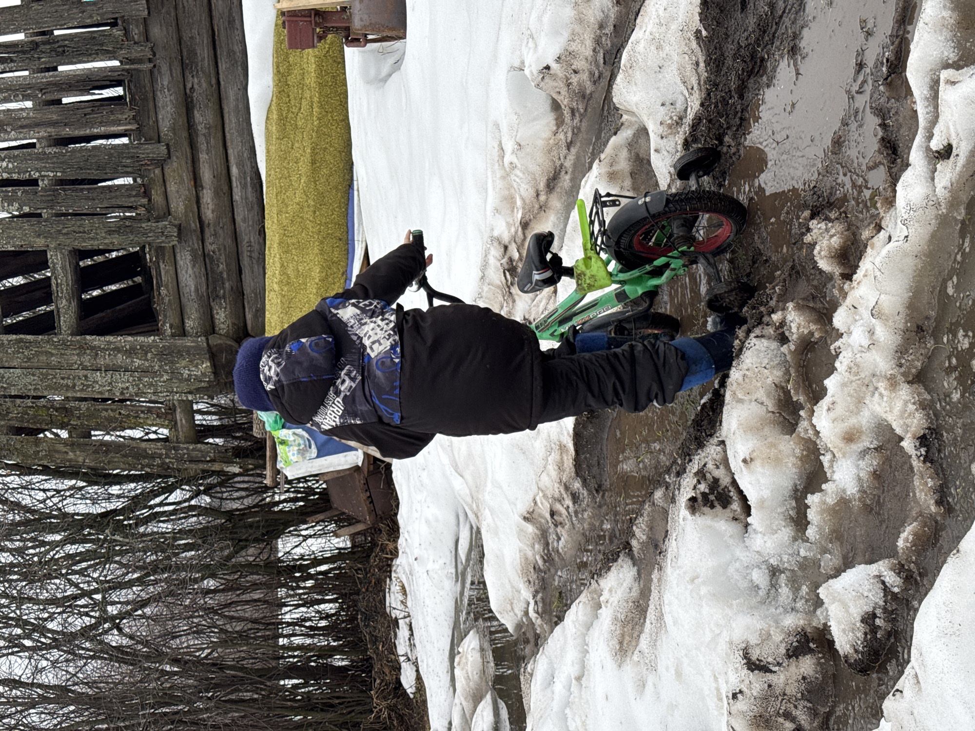 A boy stands beside a green bicycle near a snowbank, adjacent to a weathered wooden structure and bare trees. The location is possibly in Кемуль, Russia. The boy is bundled in dark clothing, including a patterned jacket and blue hat. The bicycle, which is green with red accents, has training wheels attached. The ground is a mixture of snow, mud, and water.