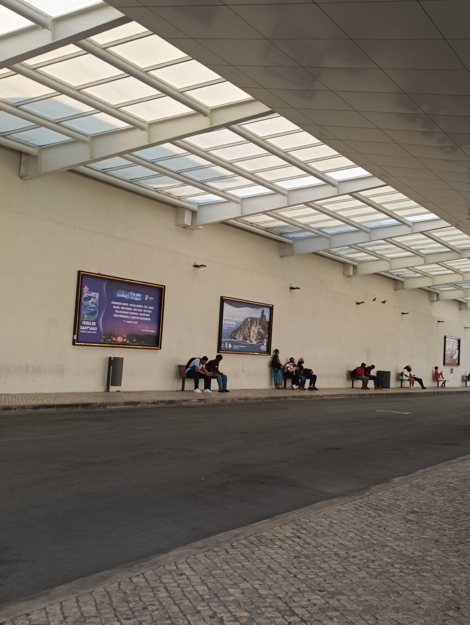 The image depicts a scene at the Setúbal Bus Terminal in Setúbal, Portugal. The terminal features a modern, angled roof with translucent panels supported by white beams, casting geometric shadows on the wall below. A long, light-colored wall spans the scene, displaying two large framed posters. The left poster advertises a "FEIRA DE SANTIAGO" (Santiago Fair) in Setúbal, dated July 25th to August 2nd, 2015. The right poster shows a scenic view of a rocky coastline and the sea.
Along the base of the wall, a paved sidewalk runs parallel to a asphalt road. Several individuals are seated on benches positioned along the sidewalk. On the left, two men are sitting on a bench, one with his back to the camera, the other facing left. Further down, a group of about five people are seated on a bench. To the right, another group of three people are on a bench, and further still, a single person is seated on a bench. A dark gray trash receptacle is located between two benches. Several small, dark objects, possibly birds, are visible on the wall in the middle ground. The overall lighting suggests it is daytime, with natural light filtering through the translucent roof panels. The scene is quiet, with people waiting on the benches.