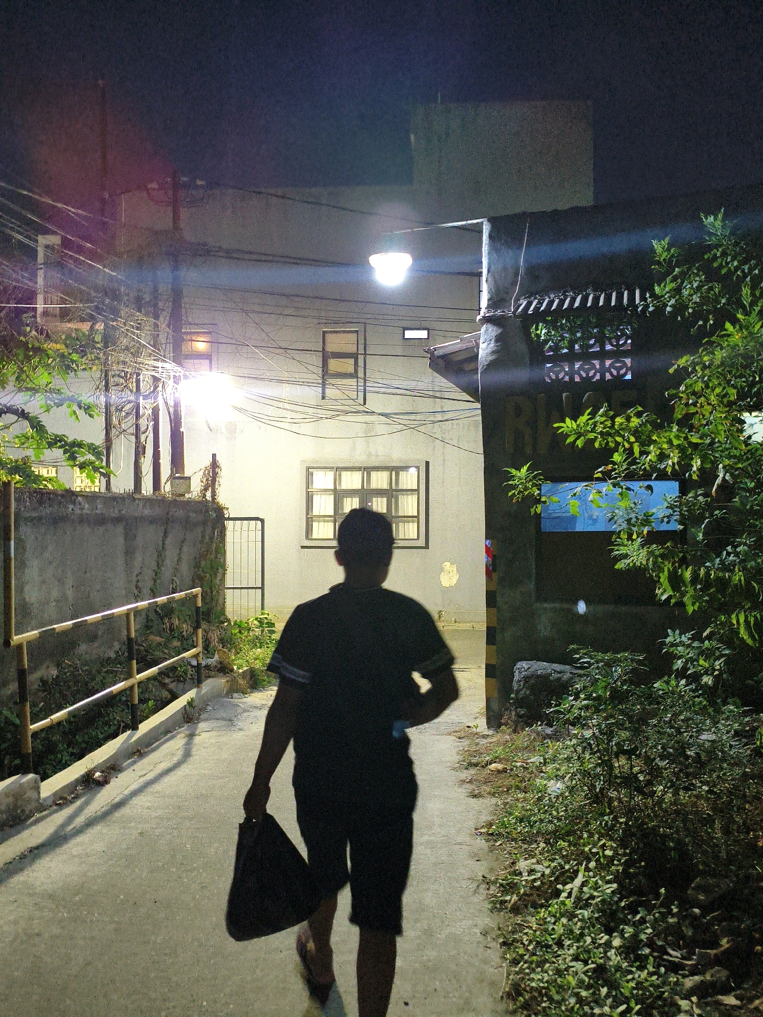 A person walks down a concrete pathway at night, carrying a dark bag in their right hand. They are silhouetted against the ambient light of street lamps and illuminated windows. The pathway is bordered by a concrete wall with a yellow and black striped metal railing on the left, and dense foliage and a dark-colored structure on the right. The background shows buildings with windows and exposed electrical wires, suggesting an urban or semi-urban setting. The lighting indicates it is nighttime, with streetlights casting long shadows. The image captures a moment of solitary movement through a dimly lit environment in the city of Bekasi, Indonesia.