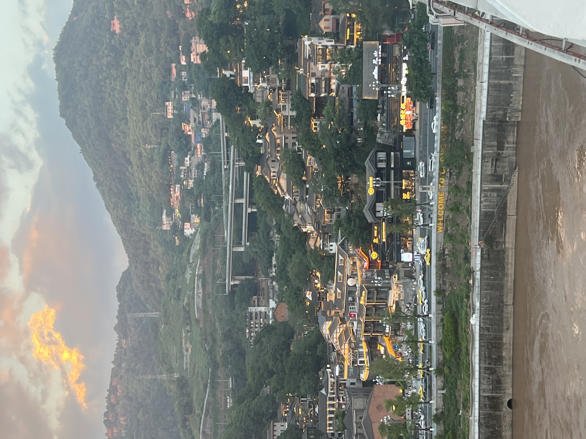 The image depicts a riverside cityscape at dusk, with buildings cascading down a verdant hillside towards a wide, murky river. The architecture is a blend of traditional styles with modern elements, featuring multi-story structures with illuminated windows and ornate facades. Many buildings are adorned with brightly lit signage, suggesting a commercial or entertainment district. A busy road runs along the riverbank, lined with traffic, including yellow taxis. A large concrete embankment separates the road from the river. In the background, a mountain covered in dense foliage rises above the urban sprawl, and an elevated highway structure is visible cutting across the landscape. The sky is overcast with dramatic clouds tinged with orange and yellow from the setting sun. The overall scene conveys a sense of vibrant urban life set against a dramatic natural backdrop. The sign "WELCOME TO CHONGQING" is visible on the embankment. The Nan'an District of Chongqing is known for its hilly terrain and riverside development, which aligns with the visual characteristics of the image.