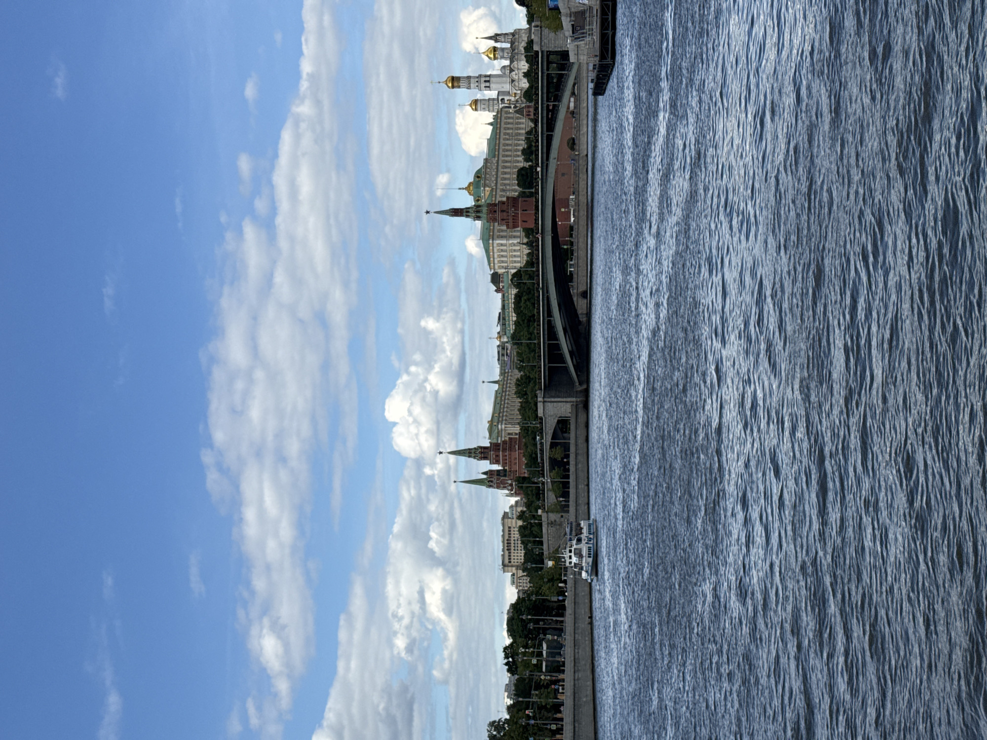 The image shows a cityscape of Moscow, Russia, next to a river. Prominent landmarks include the Moscow Kremlin with its towers and golden domes. A bridge spans the river, and boats are visible on the water. The sky is blue with scattered clouds. Buildings line the riverbank. The image is vertically oriented.