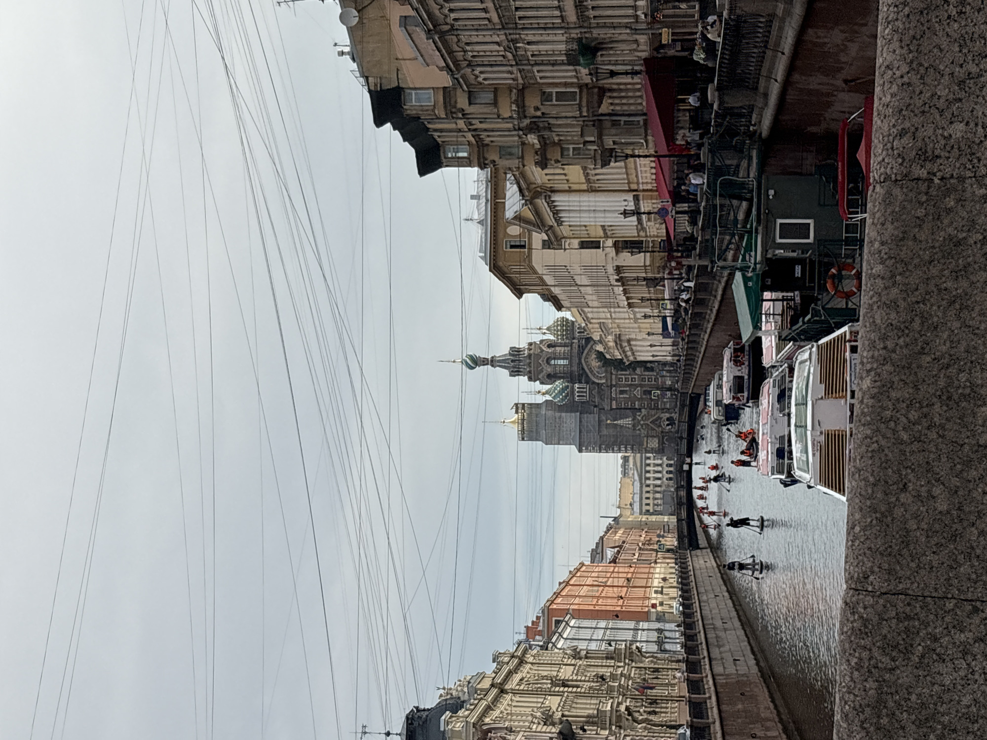 The image shows a view of the Griboyedov Canal in Saint Petersburg, Russia, with the iconic Church of the Savior on Spilled Blood visible in the background. The canal is lined with historic buildings and is filled with various watercraft, including tour boats and several people paddleboarding. The sky is overcast, and the overall atmosphere suggests a moderately busy day.
On the canal, there are at least three tour boats docked on the right side of the image, one of which is green with a red life preserver visible on its side. Several people are standing on the paddleboards, some appearing to be taking a tour or enjoying recreational activity. The buildings on either side of the canal display classic European architecture with ornate facades and multiple stories. The Church of the Savior on Spilled Blood, with its distinctive onion domes, is the dominant architectural feature in the distance. The foreground features a granite embankment of the canal. The scene is captured from an elevated vantage point, likely from a bridge. Overhead, a complex network of electrical wires crisscrosses the sky.