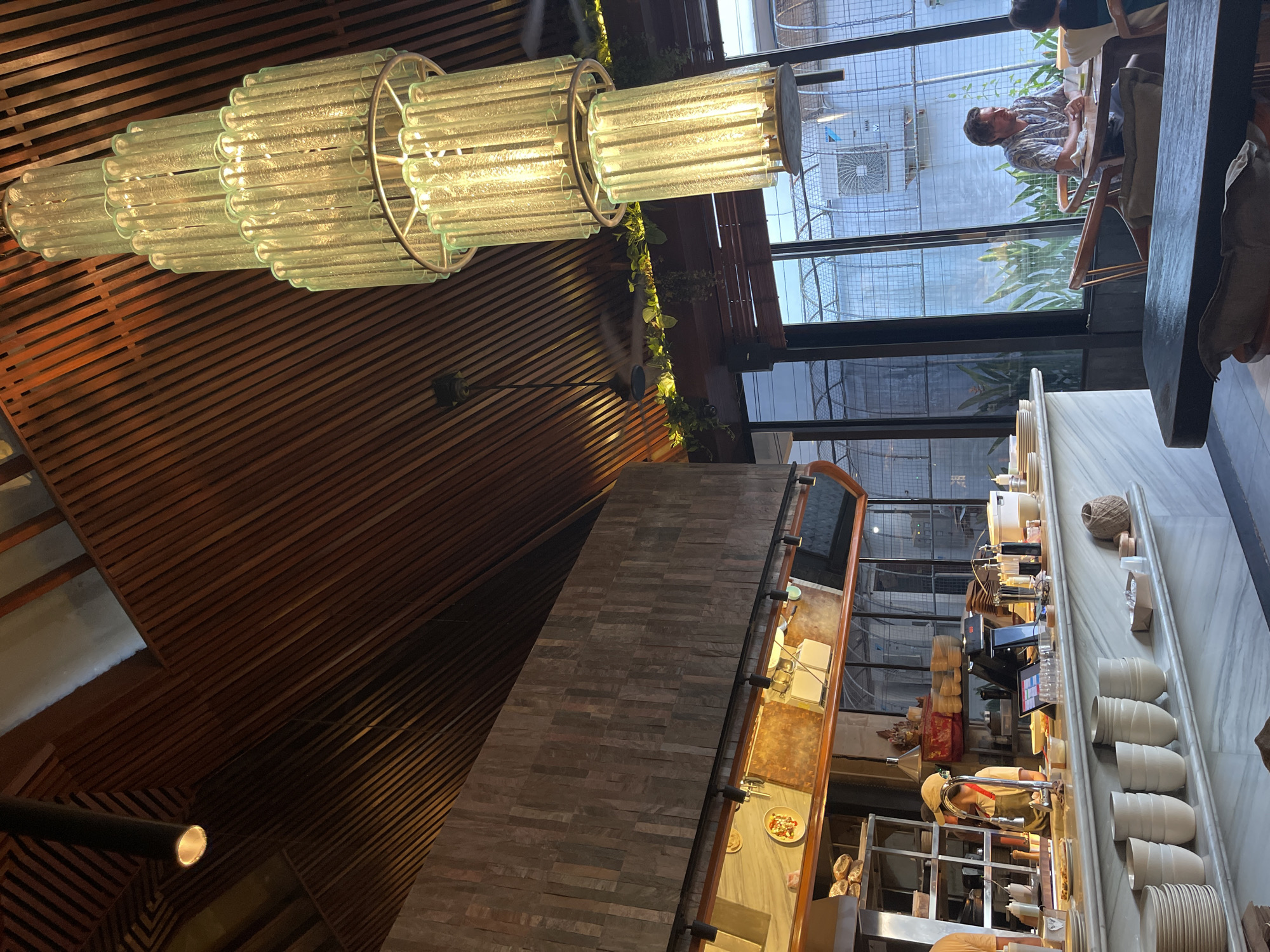 The photo shows the interior of a commercial establishment, likely a café or restaurant, in North Kuta, Indonesia. The ceiling is angled and paneled with brown wooden slats. A large, cylindrical chandelier hangs from the ceiling. A textured gray brick wall is visible.
In the background, large windows offer a view of an outdoor area. A person is sitting at a table outside. A service counter runs along the right side of the frame, featuring food preparation equipment, a cashier's system, stacks of ceramic cups, and plates. At least two employees are working behind the counter.