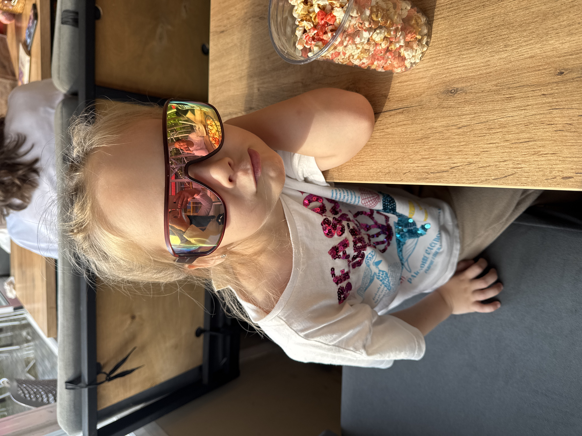 A young girl with blonde hair pulled back is seated at a wooden table, wearing oversized, reflective sunglasses and a white t-shirt adorned with colorful sequins and graphics. Her left arm is resting on the table. To her right, a clear plastic container filled with colorful popcorn is visible. In the background, a person with dark hair is seated at another table, and parts of a booth and outdoor seating area can be seen. The image appears to be taken outdoors on a sunny day, possibly at a cafe or restaurant. The scene is set in the city of Chaikovsky, Russia.