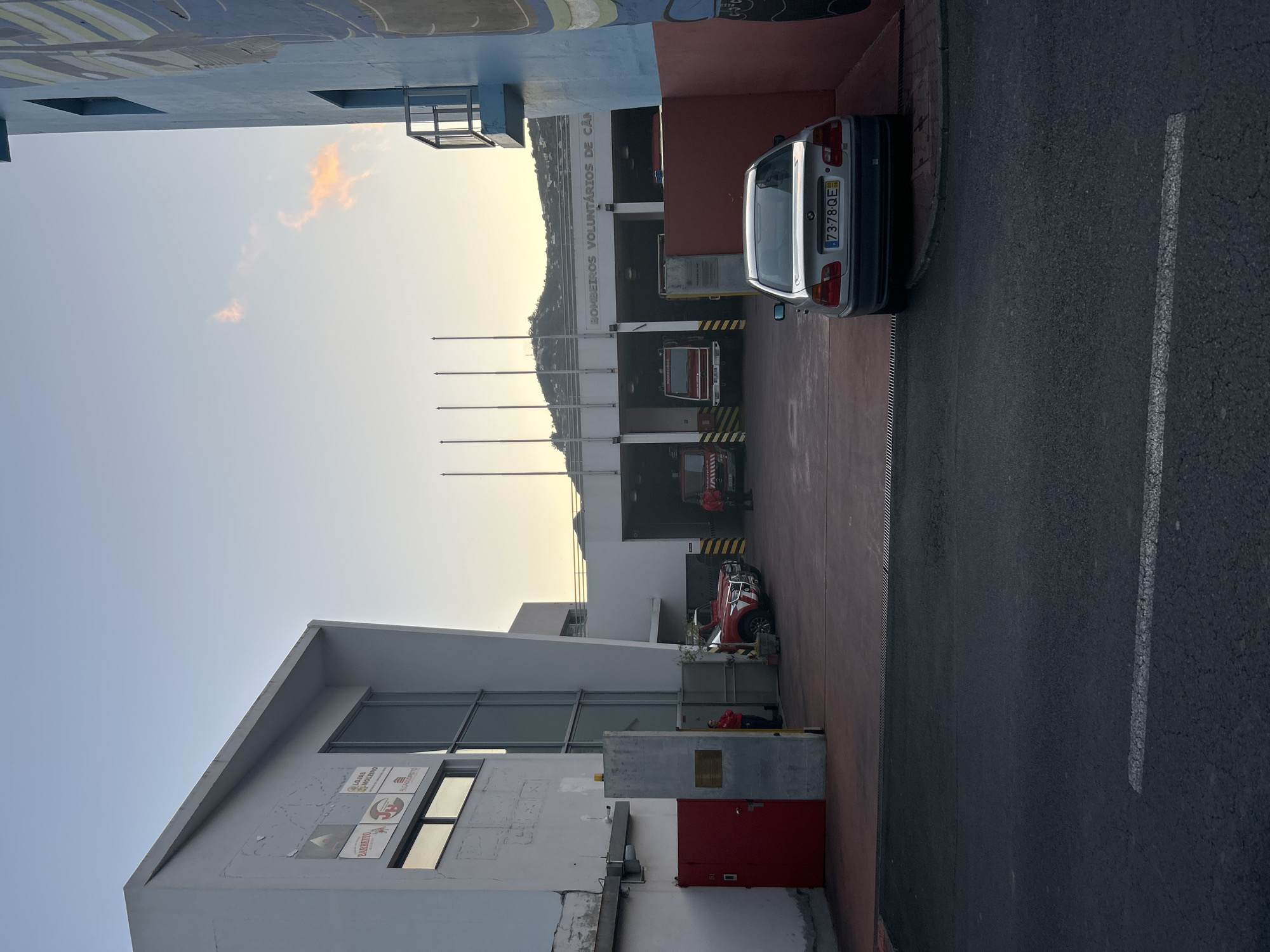 The image depicts a fire station in Câmara de Lobos, Portugal. The building has the inscription "BOMBEIROS VOLUNTÁRIOS DE CÂMARA DE LOBOS." There are multiple fire engines within the station's bays. A silver car with the license plate "73-78-QE" is parked adjacent to the station. Outside, near a white building extension with signage, a person in red is visible lying on the ground. The environment shows a street with a white line marking and a partly cloudy sky.