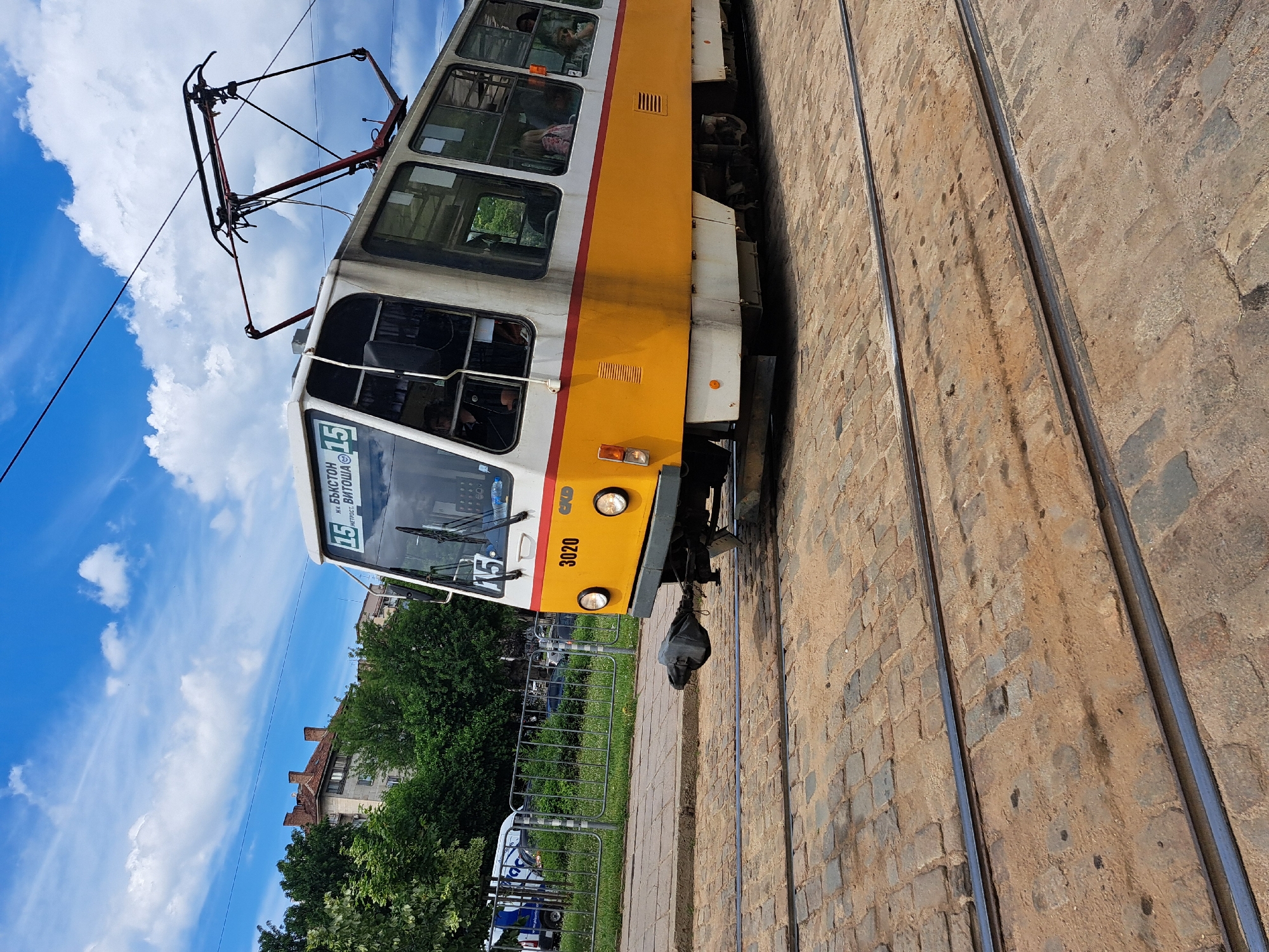 The image shows a yellow and white tram on a brick-paved street next to tram tracks. The tram has large windows, and people can be seen inside. The tram has the number "15" displayed prominently on the front, along with Cyrillic text that reads "ул. БЪКСТОН - ВИТОША". The tram's vehicle number is "3020". The scene appears to be outdoors on a sunny day, with a bright blue sky and white clouds. Trees and buildings are visible in the background, suggesting an urban setting. The perspective of the image is slightly tilted, making it appear as if the tram is leaning. The brickwork of the street and the metal tram tracks are detailed. There is a black bag hanging from the front of the tram. The image has a vibrant feel, likely taken during daylight hours.