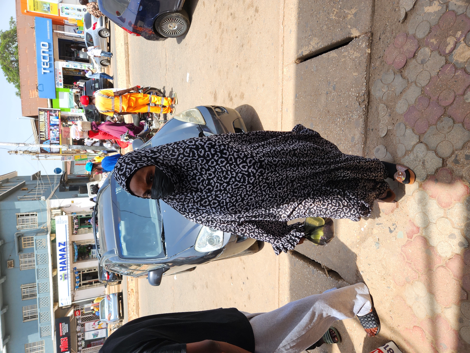 The image depicts a woman wearing a black and white patterned garment, headscarf, and a black face mask, standing on a sidewalk next to a street. She is wearing sandals. The street appears busy, with a dark grey car directly in front of her and other vehicles visible in the background. There are several businesses across the street, including one with a prominent "HAMAZ" sign. A "TECNO" sign can also be seen further down the street. The sidewalk is a mix of grey concrete and patterned pink and grey tiles. Another person's legs and part of their outfit can be seen in the lower portion of the image. The exact location is difficult to pinpoint, but appears to be an urban commercial area.