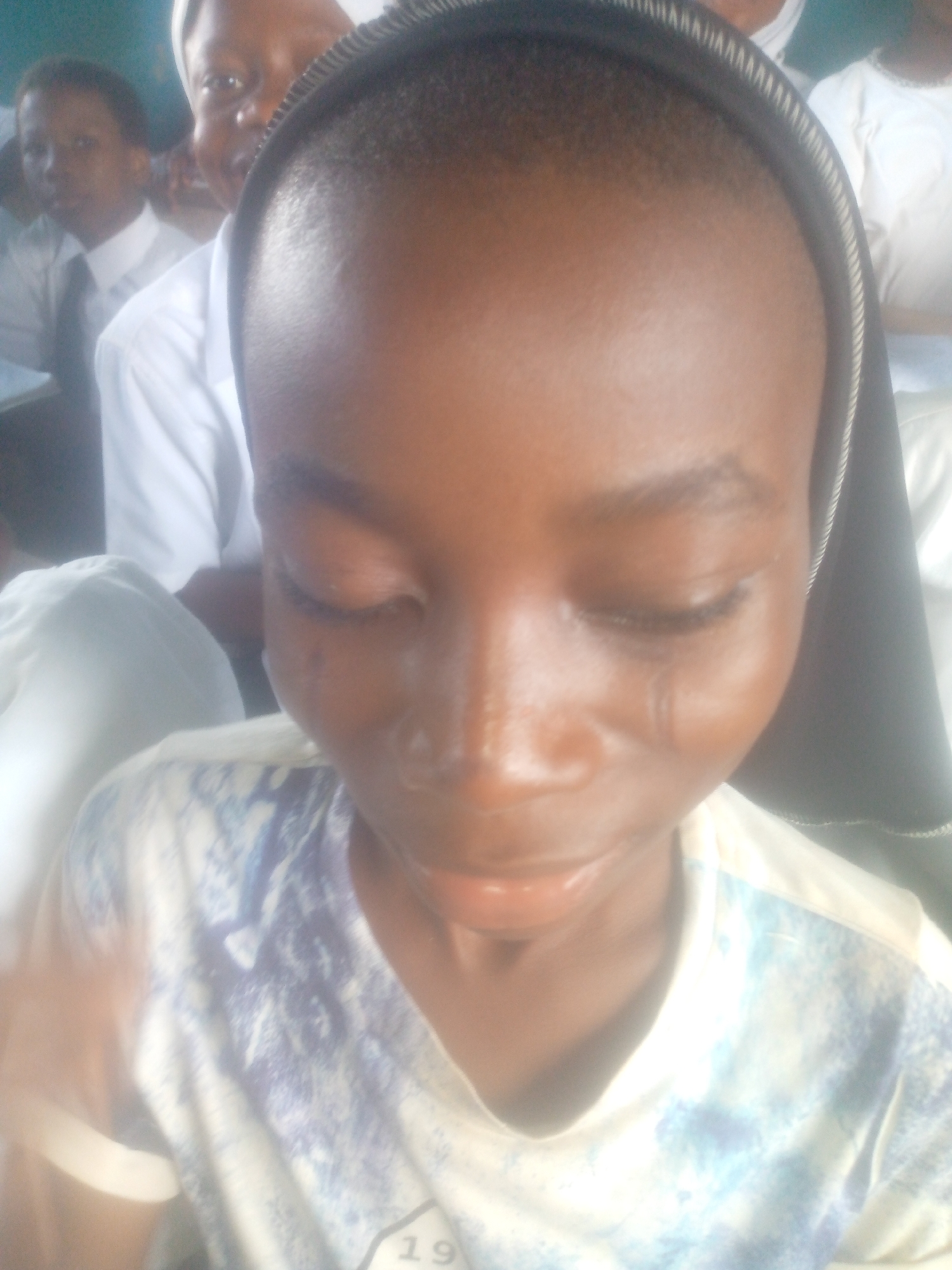 A close-up shot shows a young African person, possibly a student given the presence of uniformed classmates in the background. They are wearing a patterned shirt and have their eyes closed. Behind them, several other individuals are seen in white shirts and ties, typical of a school uniform. One person in the background also wears what appears to be a white head covering, possibly a nun's veil. A dark band is present around the subject’s head. Tears are visible on the subject's face. The location is Unknown, Unknown.