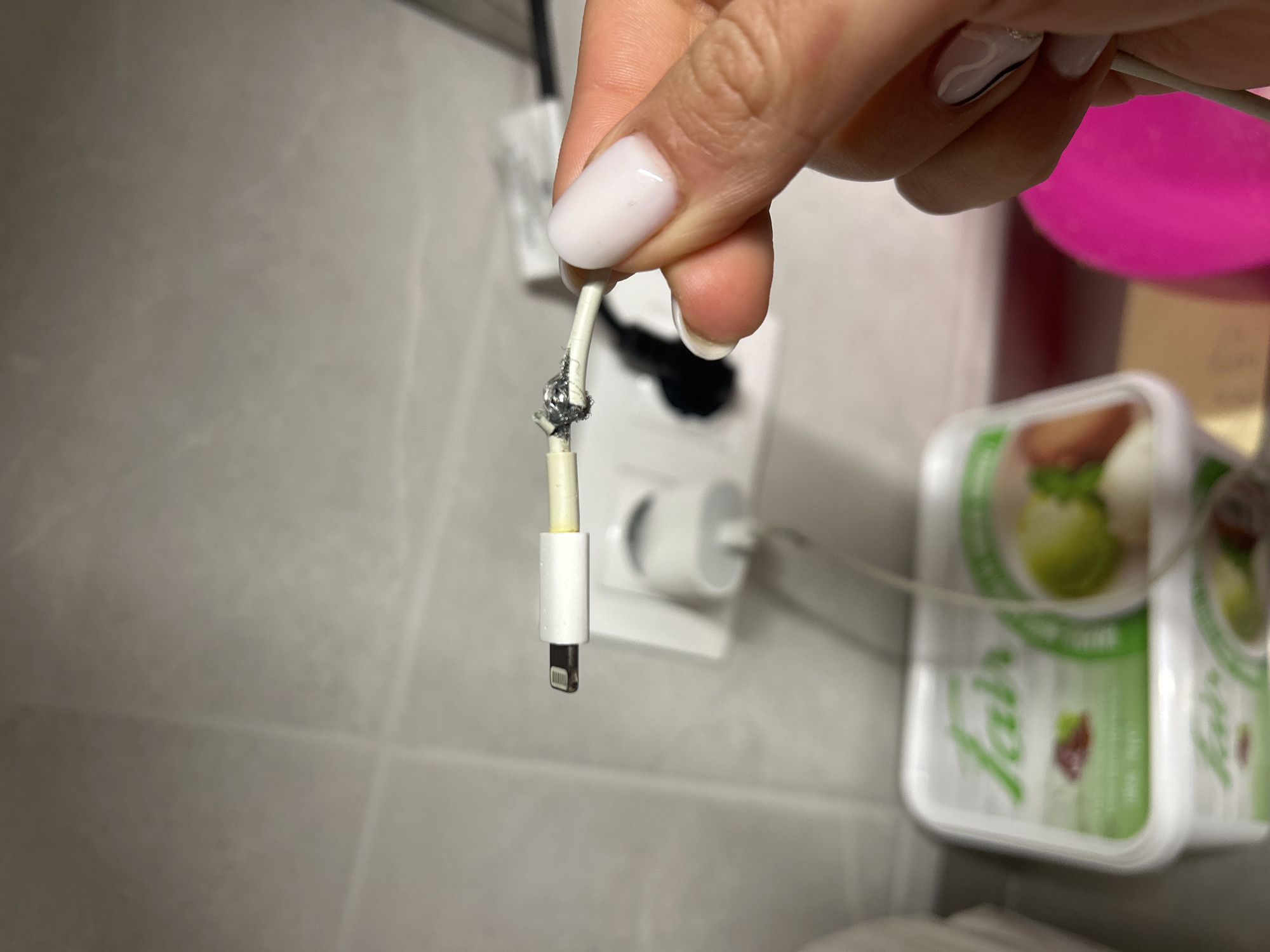 A person is holding a damaged white Lightning cable plugged into a white wall outlet, revealing visible signs of burning and exposed wiring near the connector. The background includes a tiled wall and blurred containers of "Galba" mozzarella cheese. The location appears to be indoors, possibly a bathroom or kitchen. There is no specific information to confirm the location of this scene is Burgas, Bulgaria.