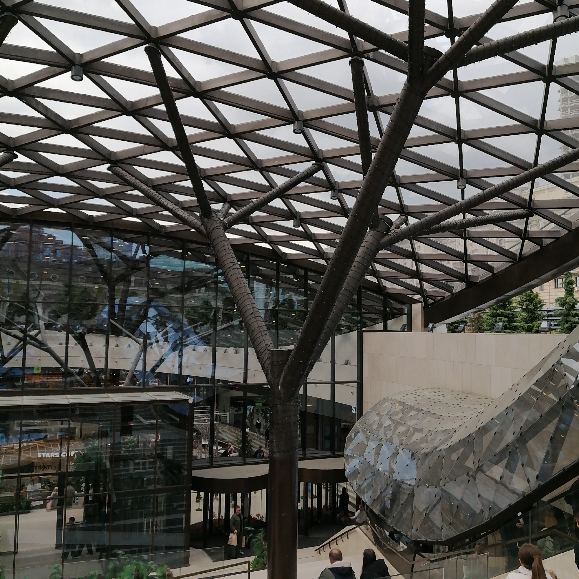 The image shows the interior of a modern building in Moscow, Russia. A key feature is the elaborate geometric glass and steel roof structure, which allows diffused light to enter the space. Large, tree-like metal supports extend from the floor to the roof.
The interior features multiple levels and various retail spaces. A "Stars Coffee" shop is visible. People are seen walking around, some on stairs. A large, amorphous geometric structure is to the right, possibly related to an escalator or stairway.
The environment is modern, clean, and well-lit. The image appears to be taken during the daytime on a cloudy day.
