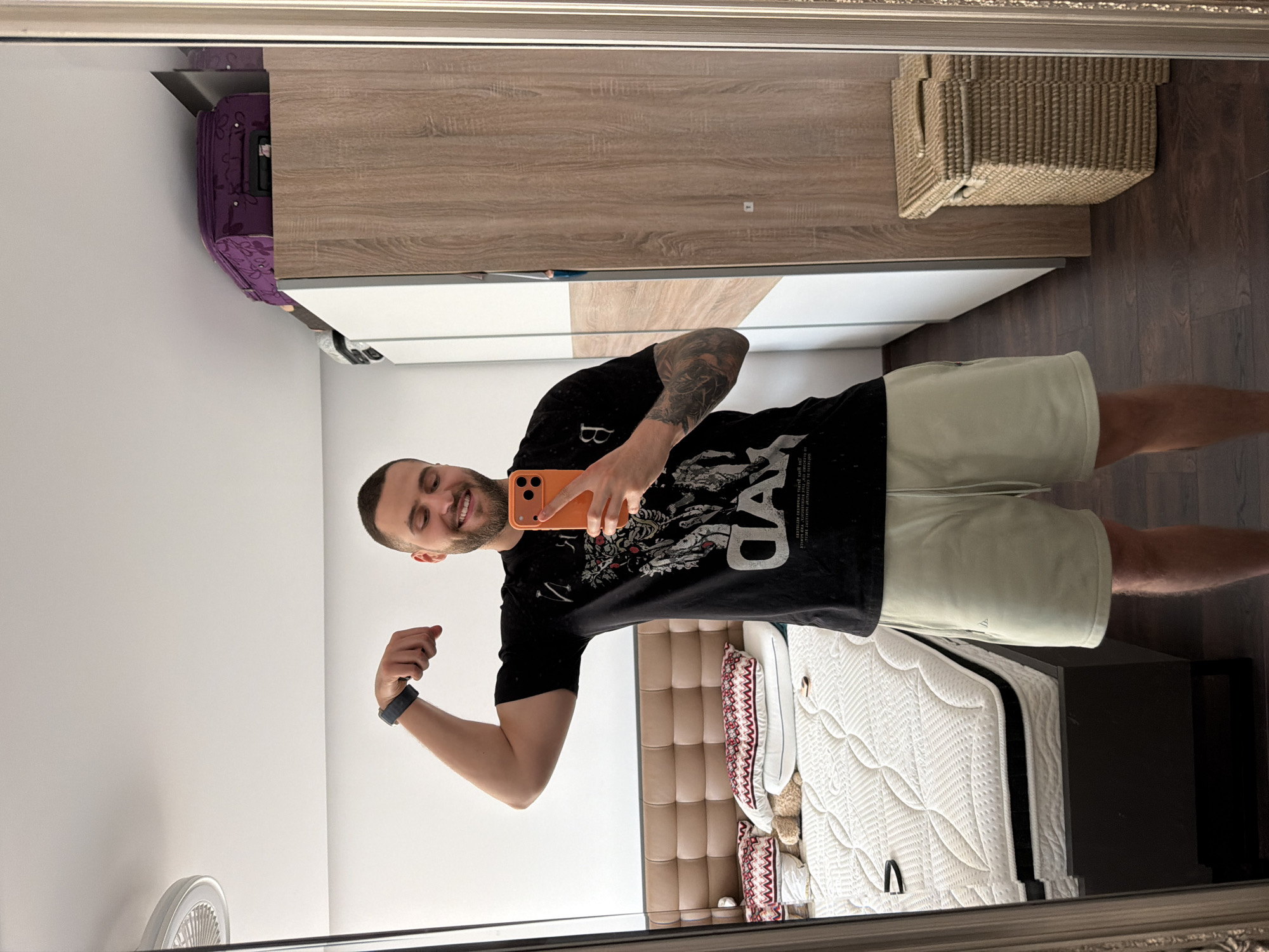 A man is taking a selfie in a mirror. He is wearing a black t-shirt, light green shorts, and a black smartwatch. He is holding an orange smartphone. He is flexing his bicep. The reflection shows a bedroom interior, including a bed, a nightstand, a wardrobe, and a basket. There is a suitcase on top of the wardrobe. The environment appears to be an apartment. The location might be in Sofia, Bulgaria.